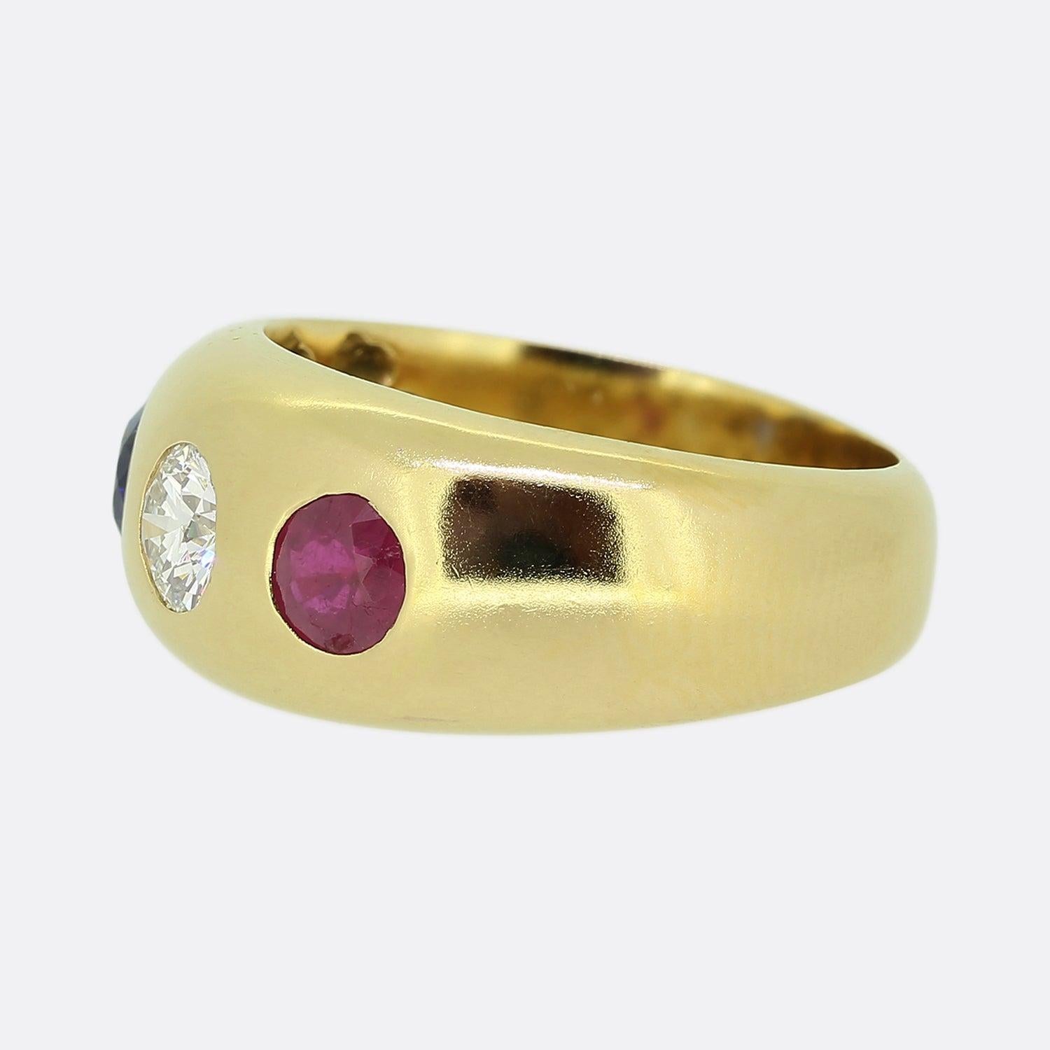 Here we have a classic three-stone gypsy ring. This piece has been crafted from 18ct yellow gold and showcases a trio of precious gemstones. A single round brilliant cut diamonds sits at the centre of the face and is flanked on one side by a round