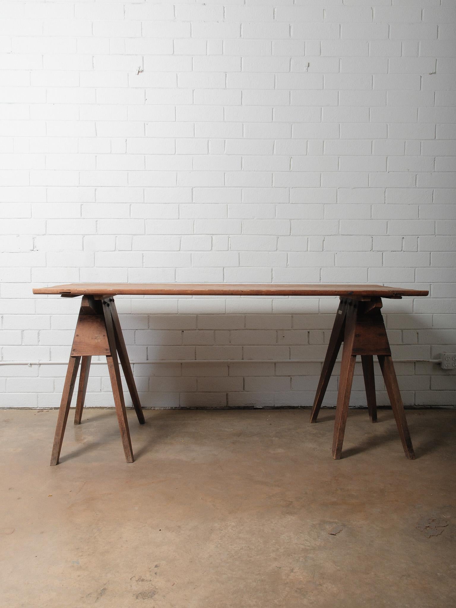 Une table de travail vintage à chevalet de scie, merveilleusement usée par le temps, avec un plateau en pin massif reposant sur une paire de bases robustes de type tréteaux. Sa construction simple et fonctionnelle incarne un design utilitaire à