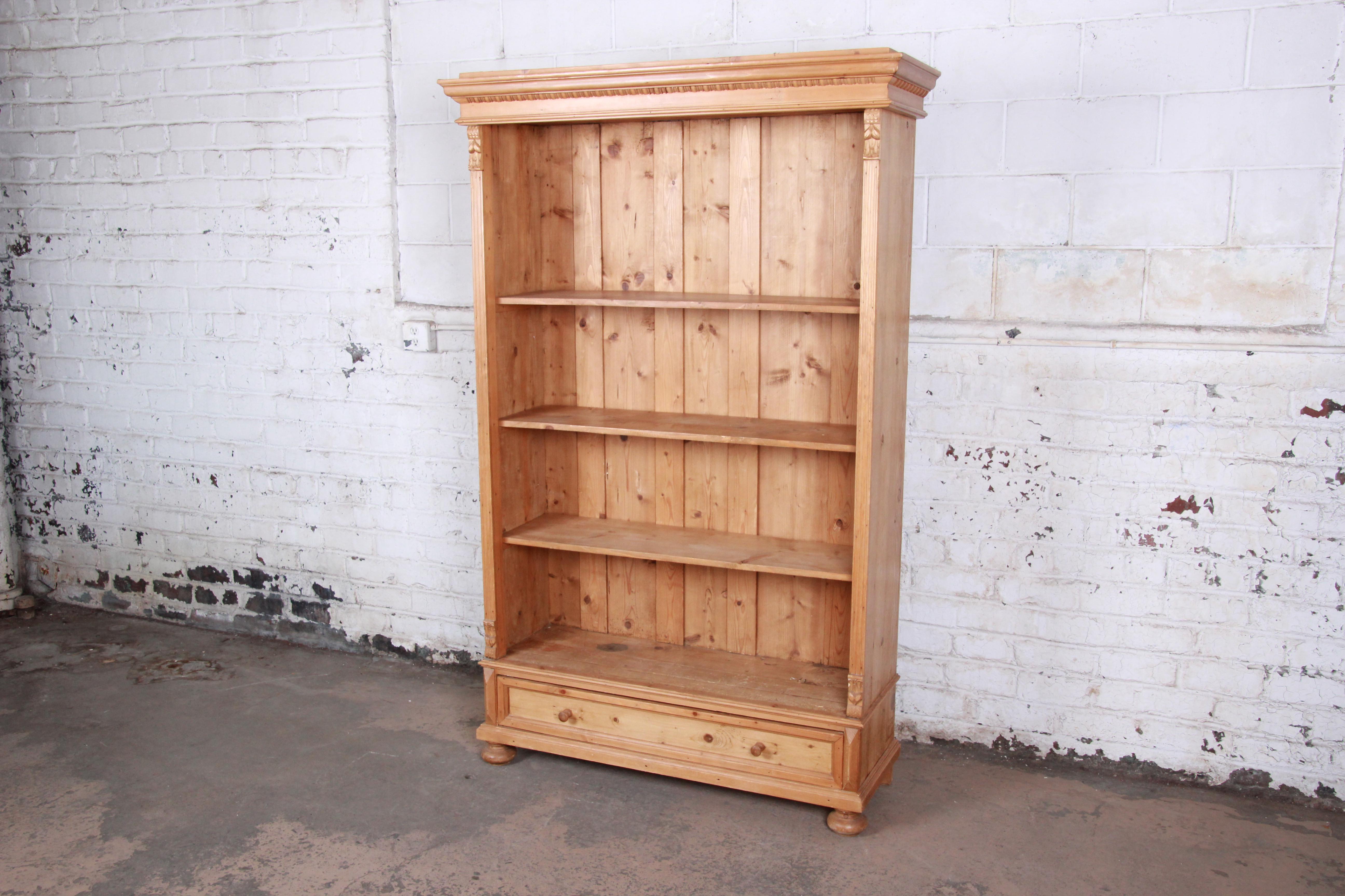 A gorgeous primitive solid pine bookcase. The bookcase features beautiful carved wood details and nice knotty pine wood grain. It offers ample room for storage and display, with three open shelves and a single dovetailed drawer below. The bookcase