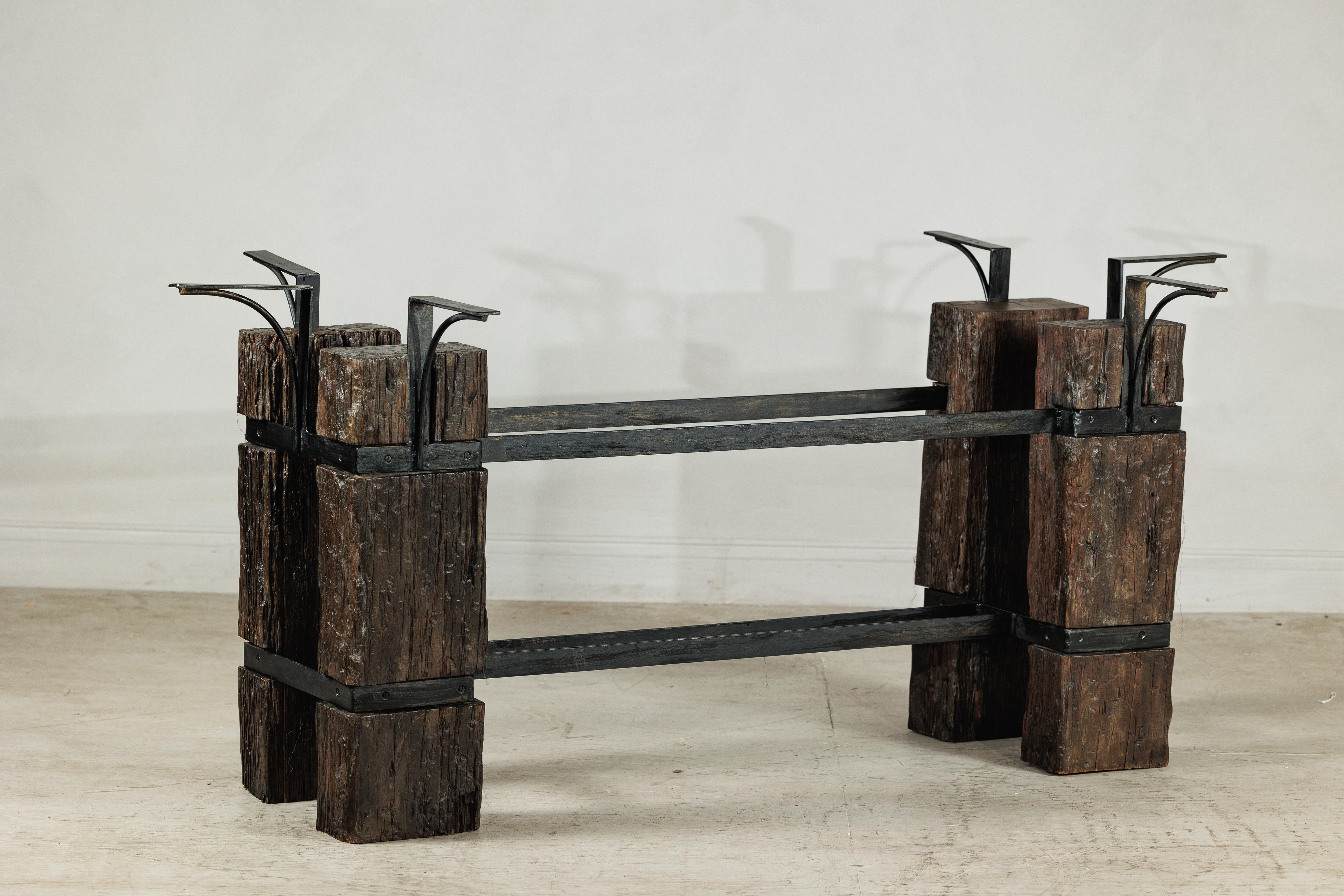 Dieser rustikale Vintage-Tischsockel kombiniert die rohe Kraft von verwittertem Holz und geschmiedetem Eisen zu einer kühnen und skulpturalen Grundlage für eine individuelle Tischplatte. Vier massive Holzpfosten, die eine strukturierte, abgenutzte