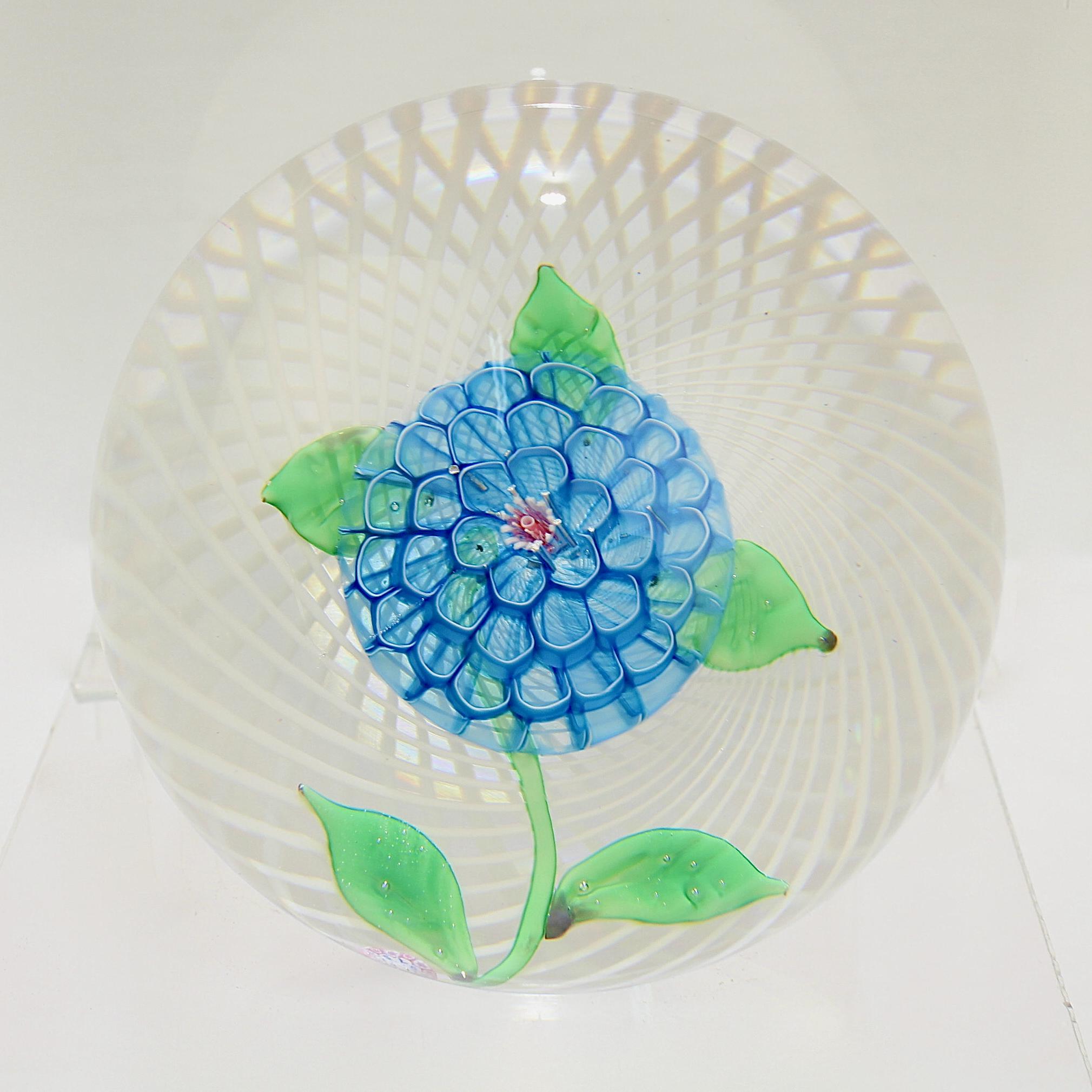 A very fine Saint Louis Camomile art glass paperweight.

It has a blue camomile (or chamomile) lamp work flower and green leaves on a white latticinio ground.

There is an SL 1975 signature cane at the base of the stem.

A very fine