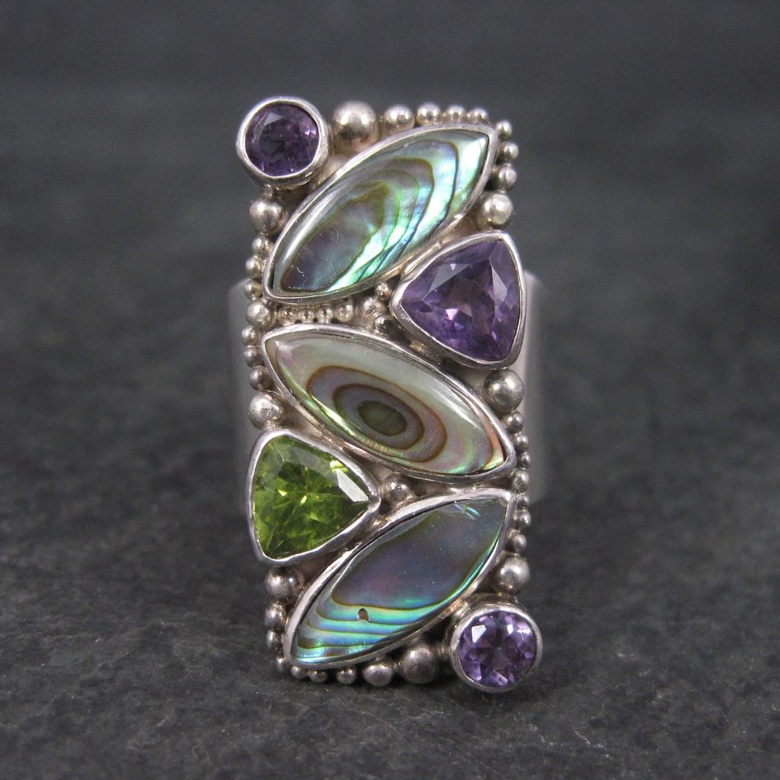 Este precioso anillo es un producto de Sajen.
Presenta concha de abulón, 1 quilate en amatistas moradas y 0,60 quilates en un único peridoto.

La cara de este anillo mide 1 1/4 pulgadas de norte a sur.
Talla: Talla como un 8 pero se ajusta como un
