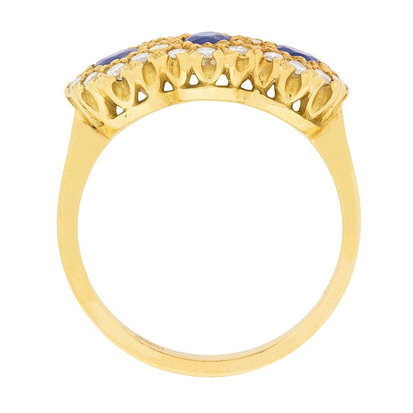 All made in 18 carat yellow gold, this ring dates back to the 1970s. There are three sapphires, each weighing 0.15 carat and they are natural. They are claw set and haloed by scalloped round brilliant diamonds totalling 0.28 carat. Estimated as G in