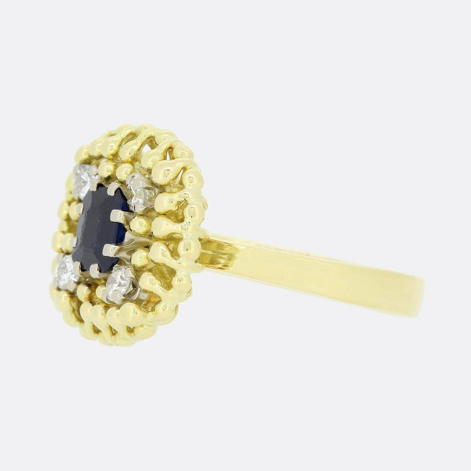 This is a beautiful vintage sapphire and diamond cluster ring. The ring features a central emerald cut blue sapphire surrounded by four round brilliant cut diamonds set on each corner of the sapphire in an abstract beaded 18ct yellow gold setting.