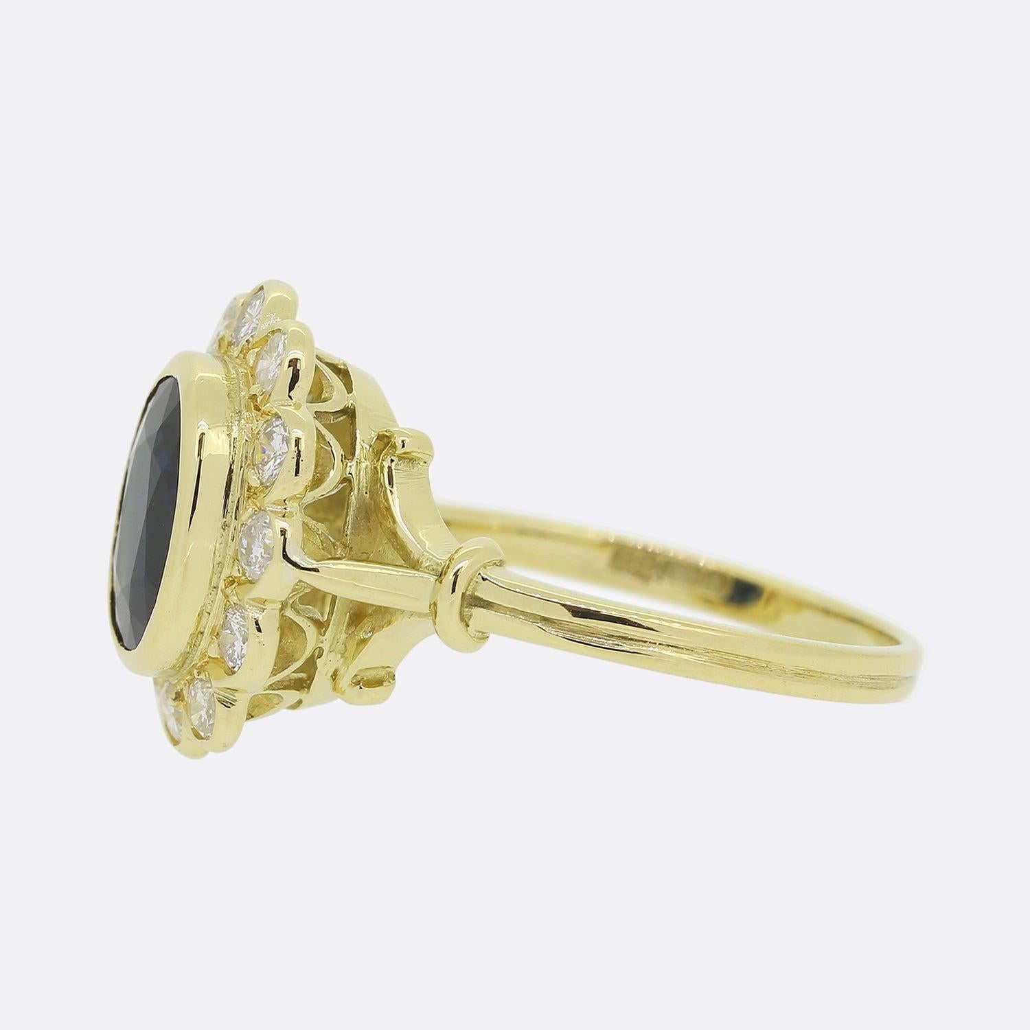 Here we have an 18ct yellow gold sapphire and diamond cluster ring. An oval shaped sapphire possessing a dark navy blue tone sits slightly risen in a rub-over setting at the centre of the face. This proud principle stone is then circulated and