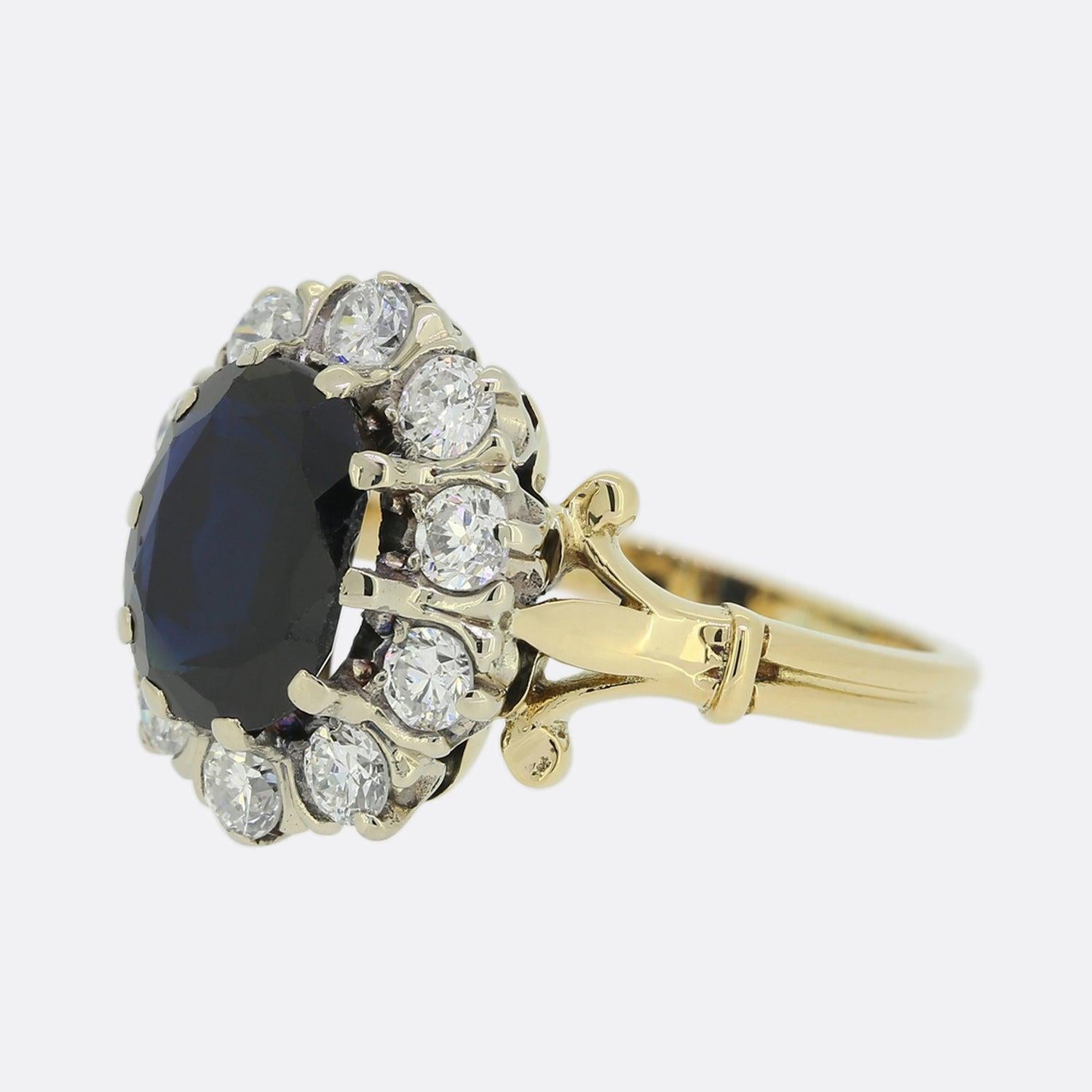 Here we have a sizeable sapphire and diamond cluster ring. At the centre of this vintage piece we find a flat, widely spread oval shaped sapphire possessing a deep royal blue colour tone. This focal stone has been set in platinum and is circulated