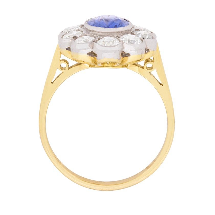 A scalloped halo of ten glittering old cut diamonds totalling one carat envelops a 1.50 carat, periwinkle blue, oval-shaped natural sapphire, all set in platinum with the balance of the ring in 18 carat yellow gold to create this 1960s rendition of