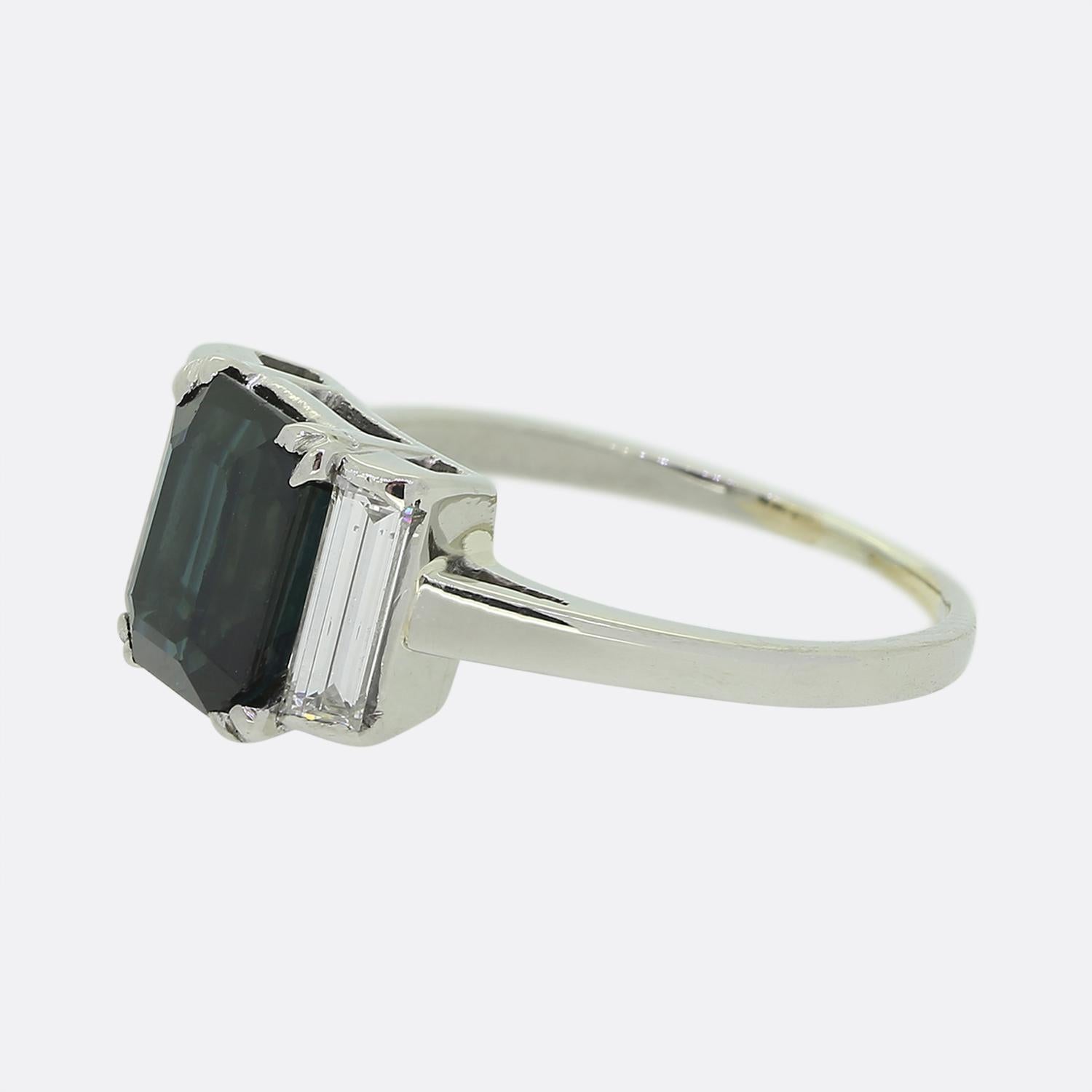 Here we have a wonderful vintage ring crafted in platinum. The ring features a dark blue central sapphire with a bright white baguette cut diamond on each shoulder. The dark blue of the sapphire is highly complimented by the contrasting bright white