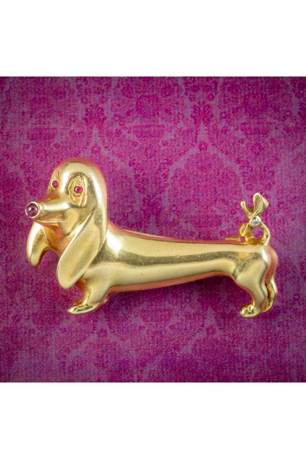 A darling vintage sausage dog brooch modelled in solid 18ct gold which has developed a lovely smoothness with age. He has a delightful and exaggerated design, with big floppy ears, a slender body and pointed nose, set with ruby eyes, nose and a