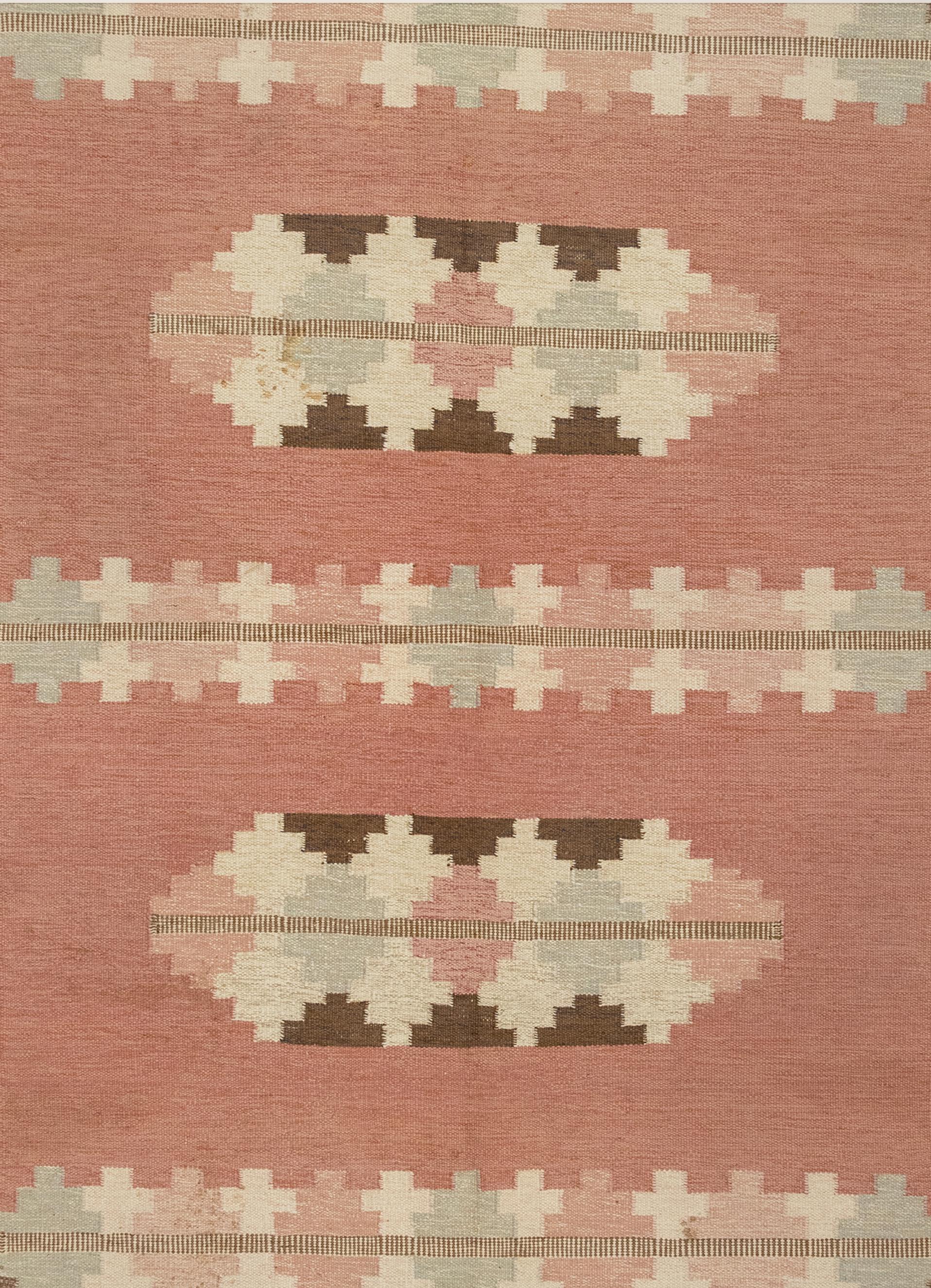 This is a Scandinavian flat-woven rug from the mid-20th century. It is signed by the designer. The color palette comprises of gentle shades of pink, icy blue, ivory, and brown in complementary geometric shapes that alternate in bands. All the