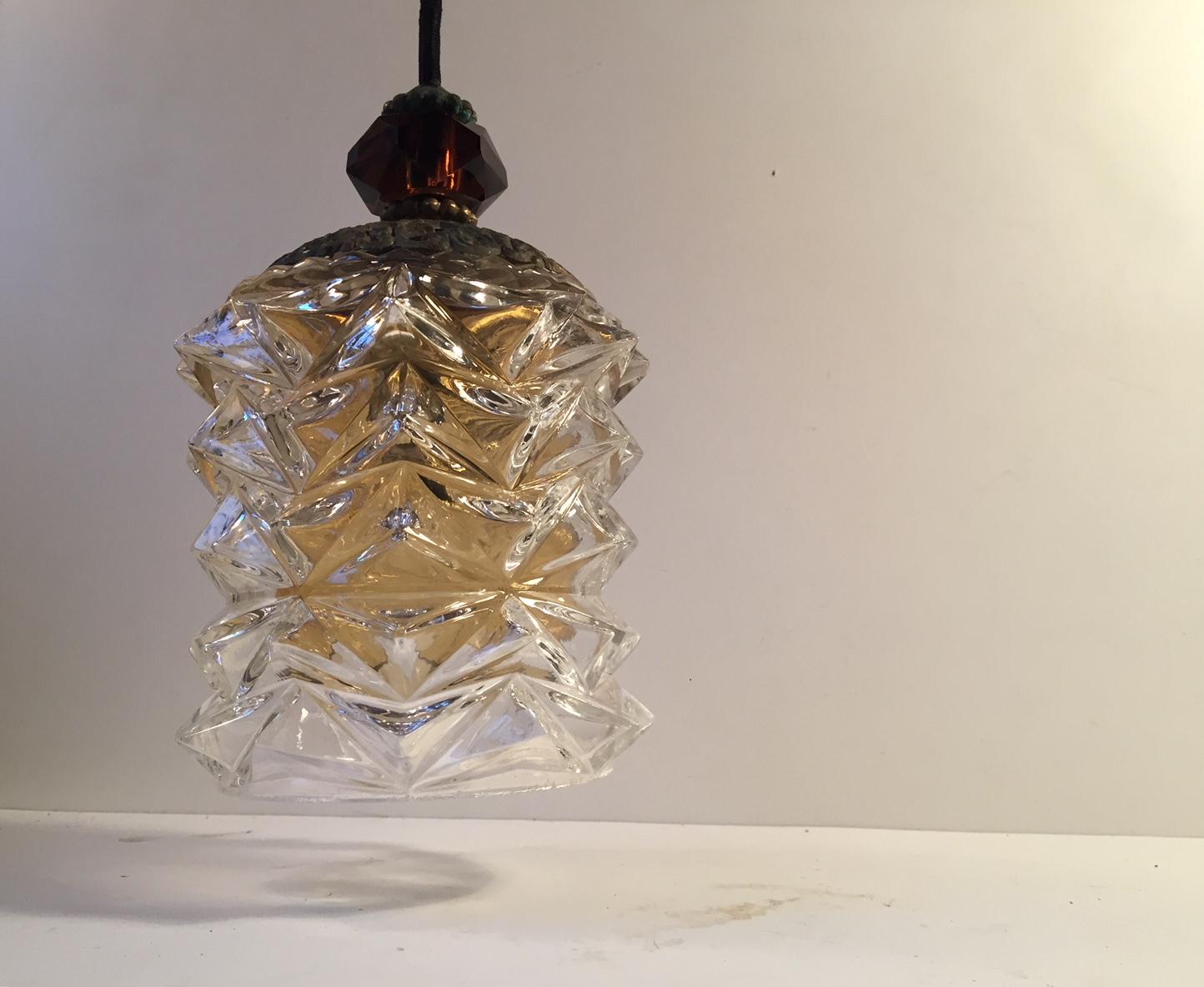 A spiky Scandinavian Modern crystal pendant light that creates a sunburst motif on the surface beneath it once lid. It features an amber glass inner shade and a amber colored faceted crystal top piece above a perforated and patinated brass sock. It