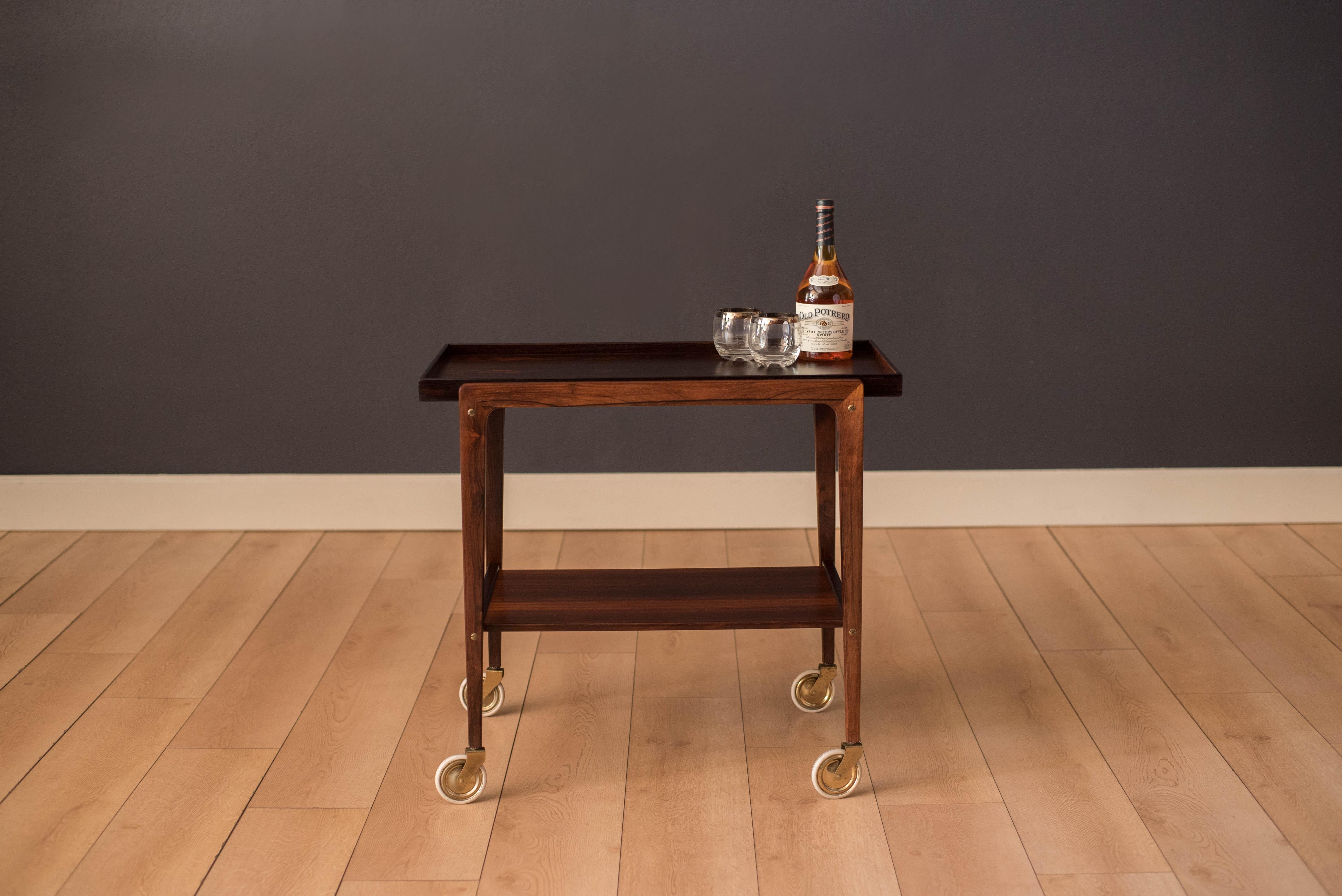 Mid-Century Modern bar cart in rosewood, circa 1960s. Features a removable tray top with dovetail construction supported by a sculptural frame with exposed bridal joints. This piece includes two-tier storage and rolls on original casters for easy