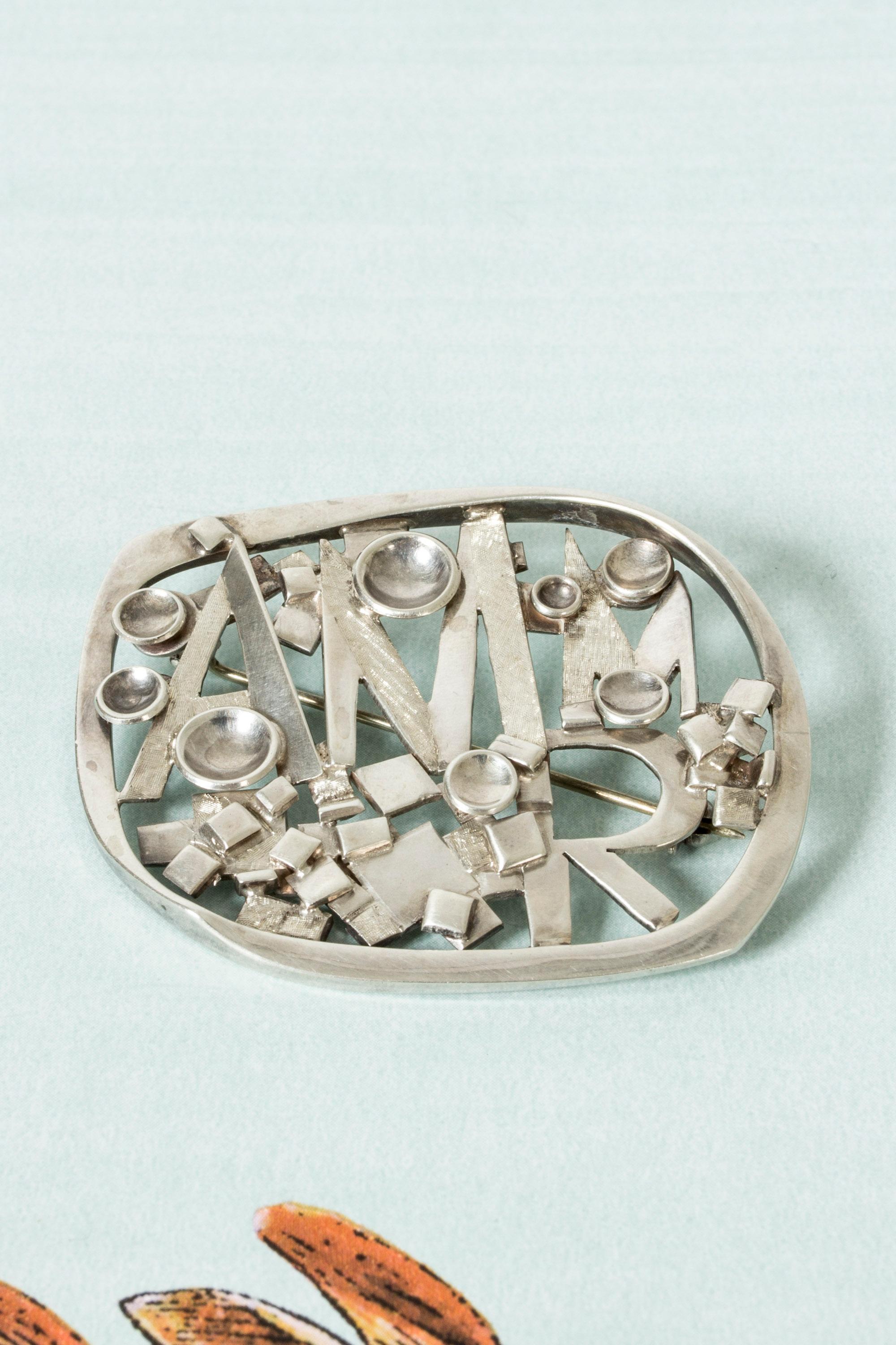 Striking silver brooch by Claës Giertta, in a cool, brutalist design. Letters and different forms make up the motif, the surfaces have different finishes that create an expressive play with light.