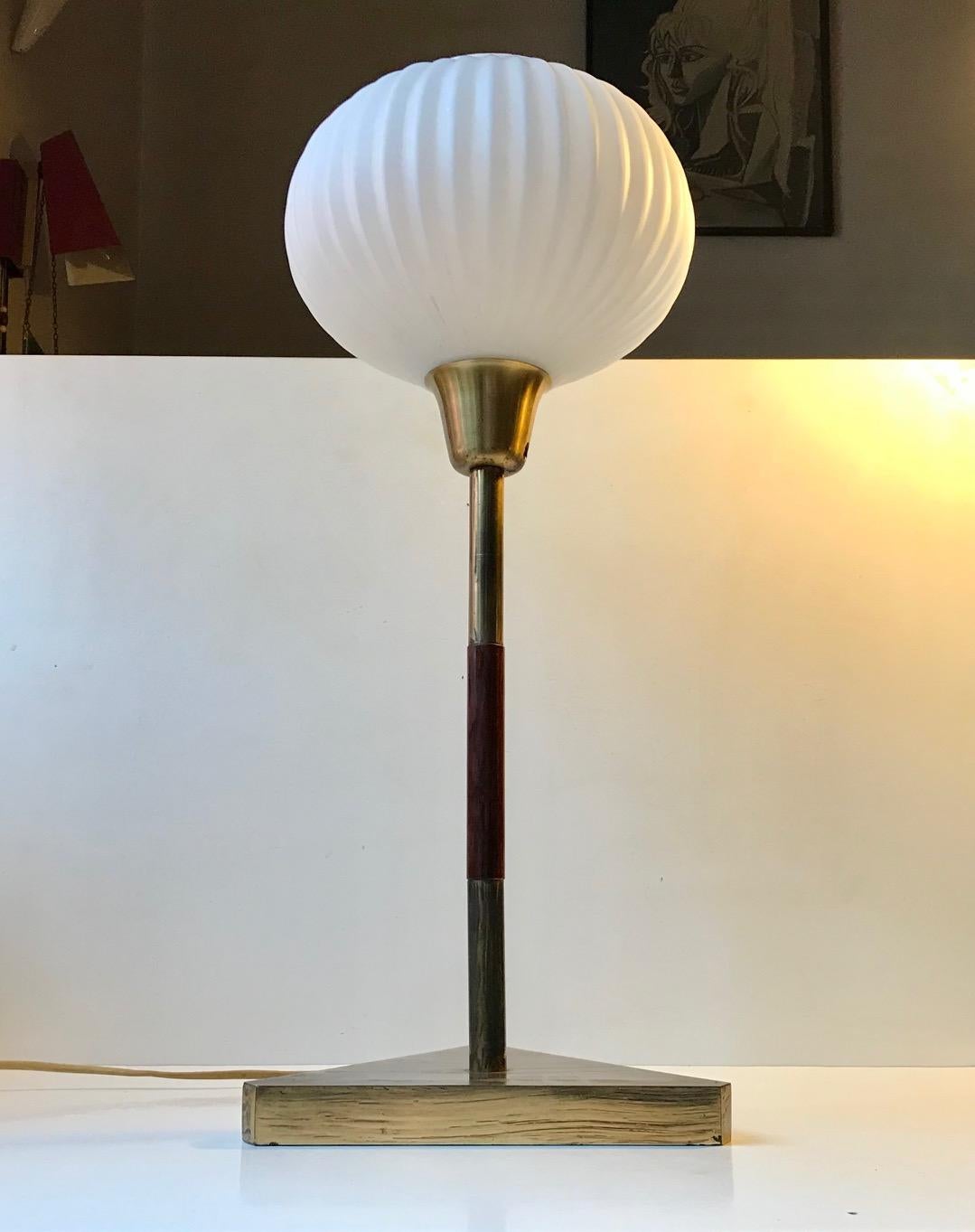 Tall simplistically composed table lamp in brass and mahogany. Mounted with a fluted spherical opaline glass shade. It features a triangular base that emphasizes the clean and well balanced Art Deco lines. It was manufactured and designed