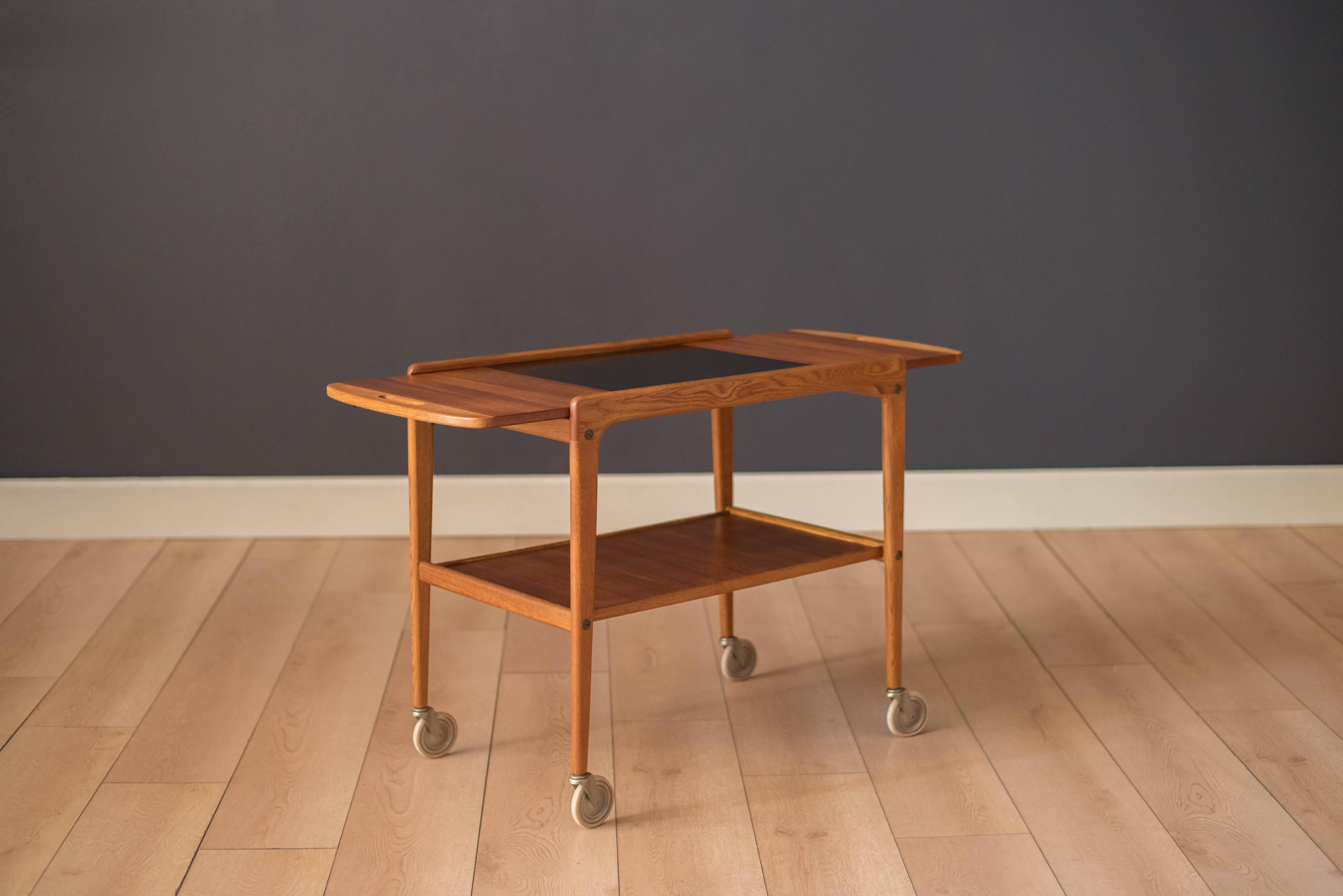 Mid-Century Modern tea or trolley bar cart in teak and oak designed by Yngve Ekstrom, Sweden. This versatile piece is equipped with a drop leaf that slides to extend for more serving space. Features an easy to maintain black laminate top and two