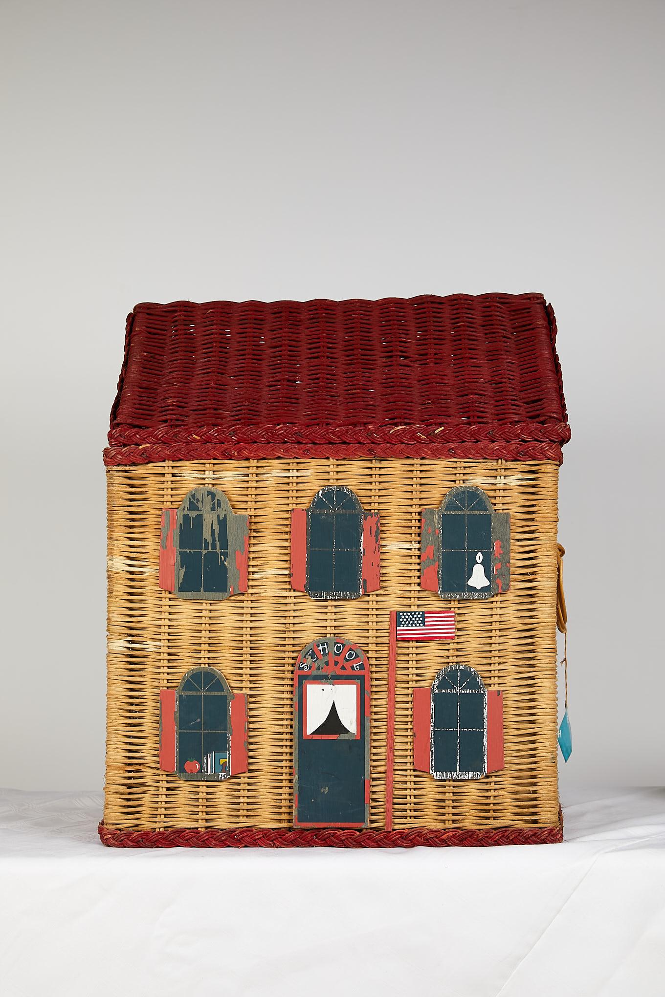 Nostalgic vintage handmade wicker toy basket in the shape of an American Colonial schoolhouse. The Classic red roof top opens on hinges to reveal storage inside and round woven handles adorn each side. The front is embellished with stylized