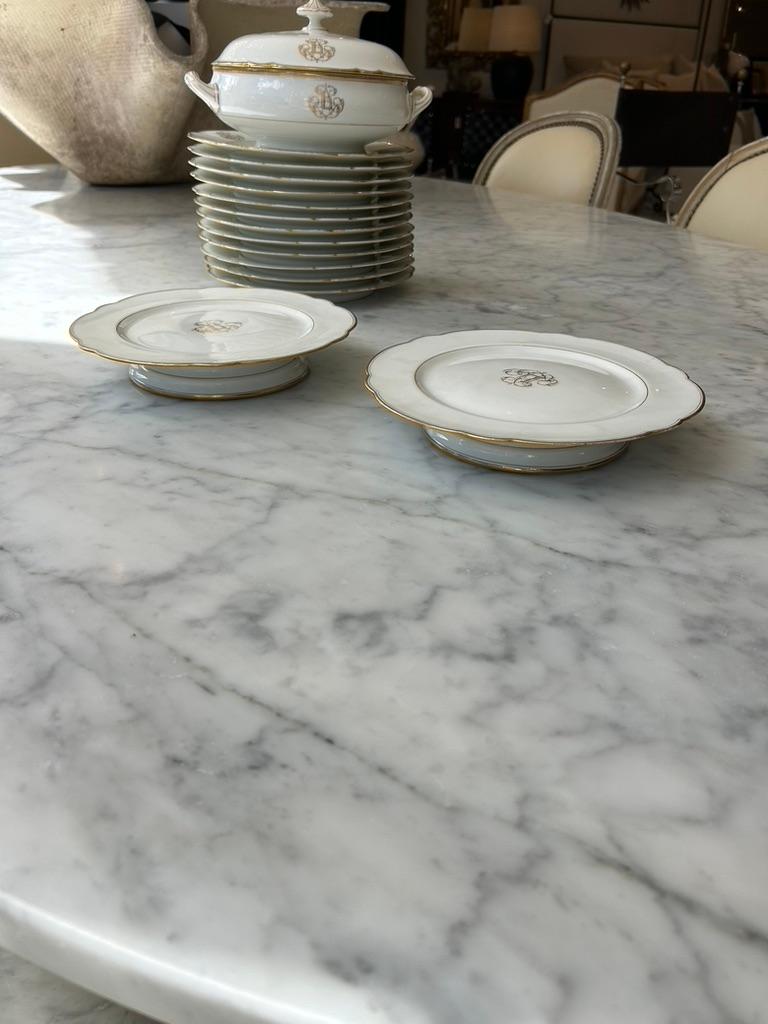 This is a set of elegant white porcelain china with gold trim detailing. The pieces include serving dishes, plates, and cups, featuring a monogrammed or crested motif in gold. The design has a classic, timeless appeal, suggesting it may be fine