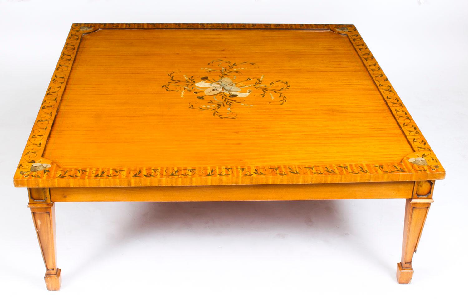 Il s'agit d'une grande et superbe table basse en bois de satin de style néo-Weraton, datant du milieu du 20e siècle.

Le plateau de table carré présente un décor peint à la manière d'Angelica Kauffman, mettant en scène des trophées et des