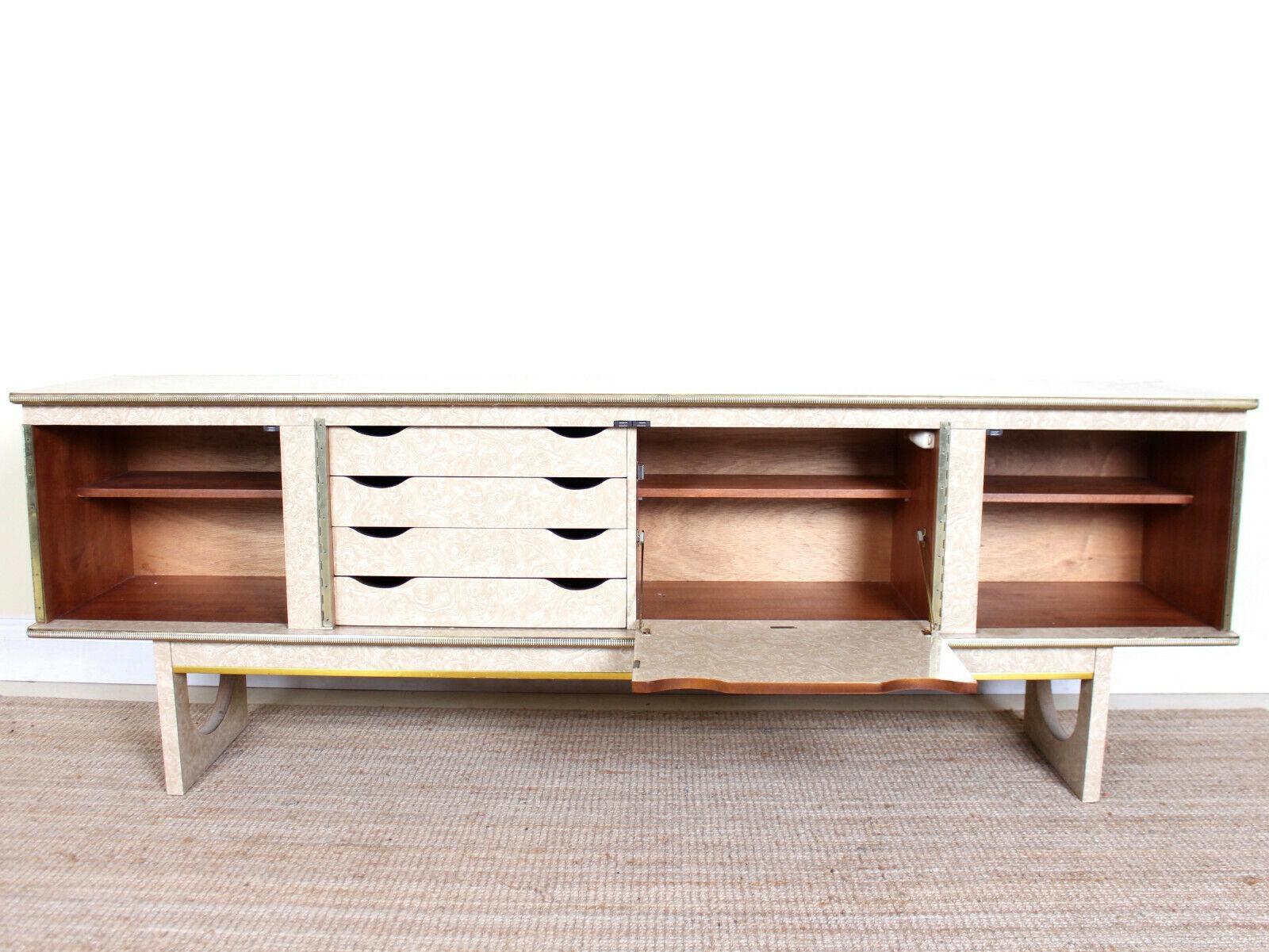 An impressive mid-20th century cocktail sideboard.
Brass trimmed bleached walnut print panels with a column of graduated trays flanked by a fall flap doors and cupboards enclosed shelving.