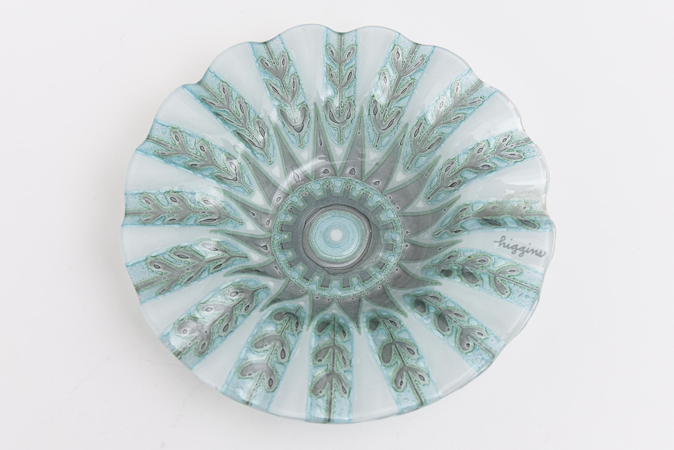 This beautiful signed mid century modern Higgins fused glass bowl has subtle colors of blue green, light turquoise and a gray silver with forms of ferns and foliage. it has a ruffled dimpled form with designs of a repeated leaf throughout. Signed