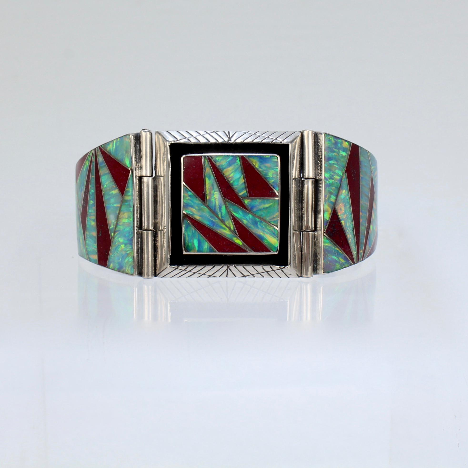 A vintage, artist signed old pawn bracelet.

In sterling silver and set sharp-angled, geometrically cut cultured opal and red jasper inlaid sections.

Hinged to the top and clasp.

Signed with what we believe is a Zuni pictograph 'sunrise'