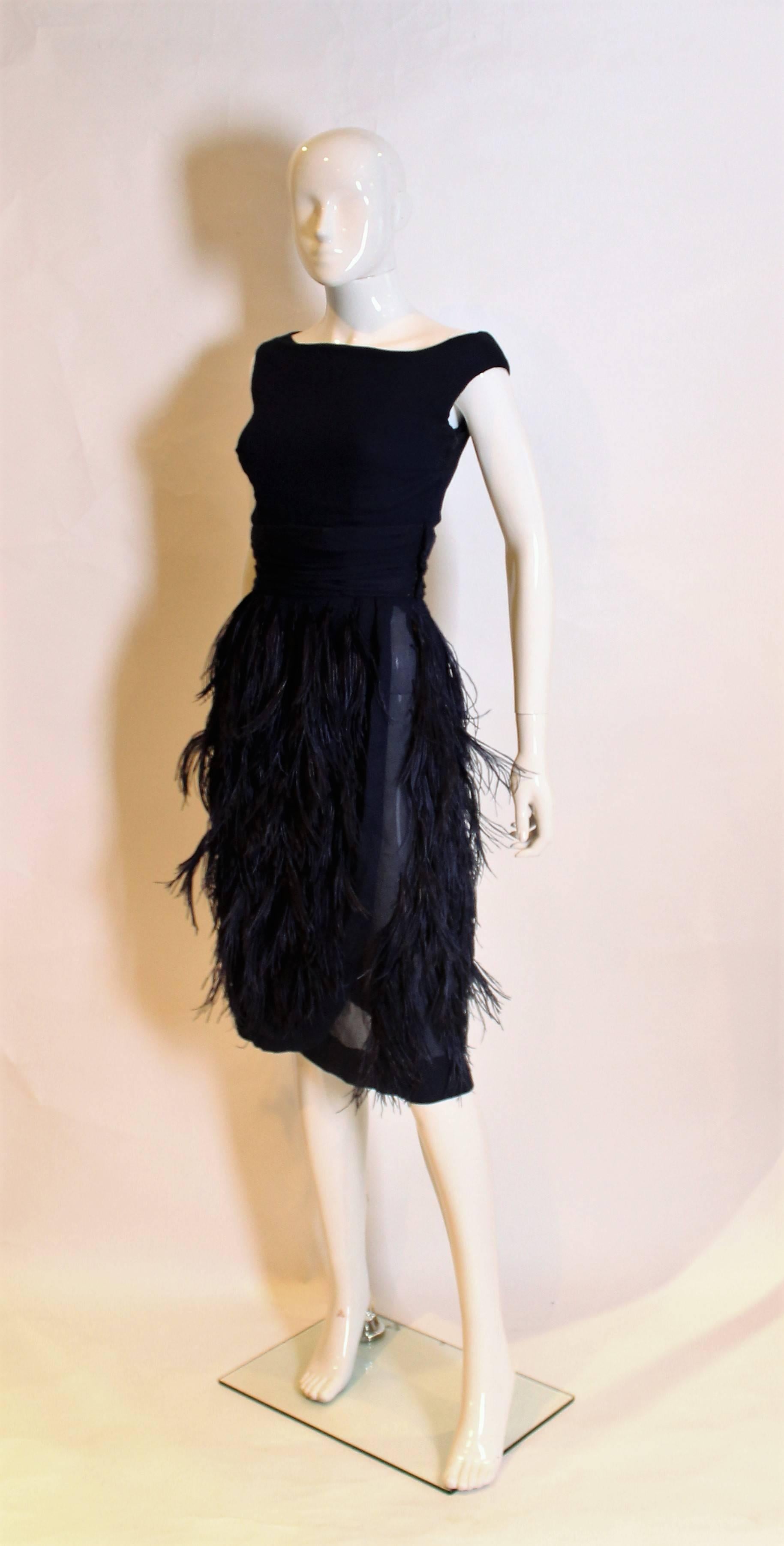 A charming and chic cocktail dress by British designer Norman Hartnell.
In a bue silk chiffon , the dress dress is sleaveless with a slip over top, and self fabric belt.There is a side zip and the skirt is covered in blue feathers.