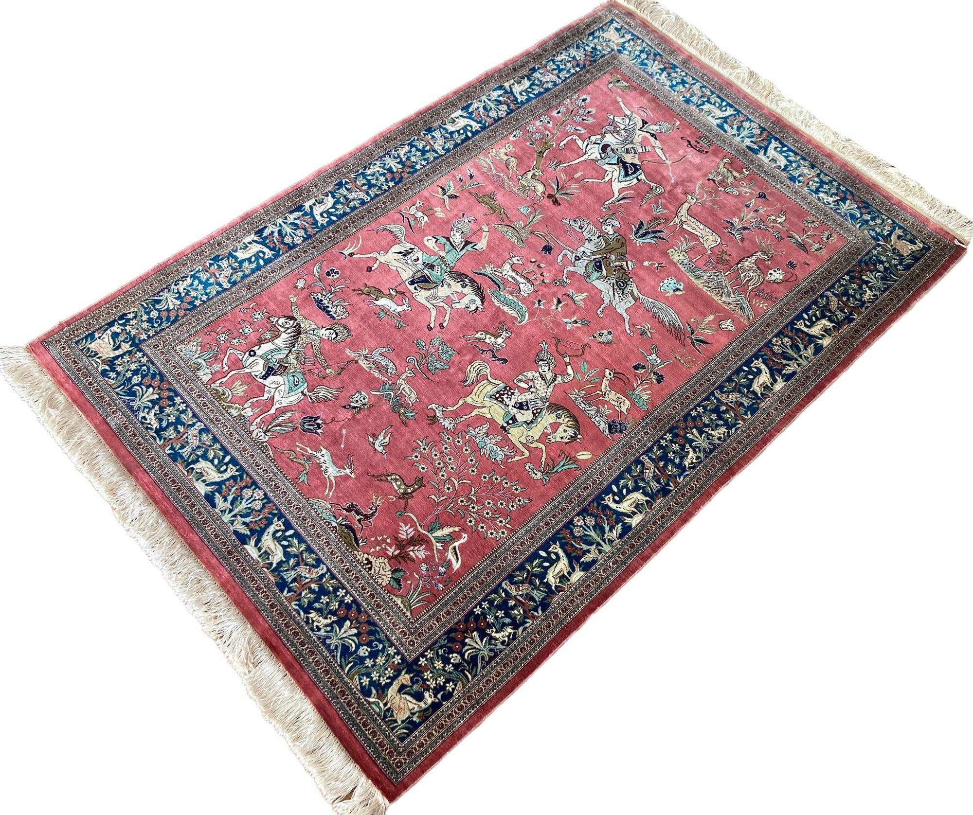A very fine silk Qum rug, handwoven circa 1980 with a traditional hunting scene on a rose field and blue border. Exceptionally fine with 10x10 knots per centimetre. The figures are particularly interesting with the saddles and jackets having