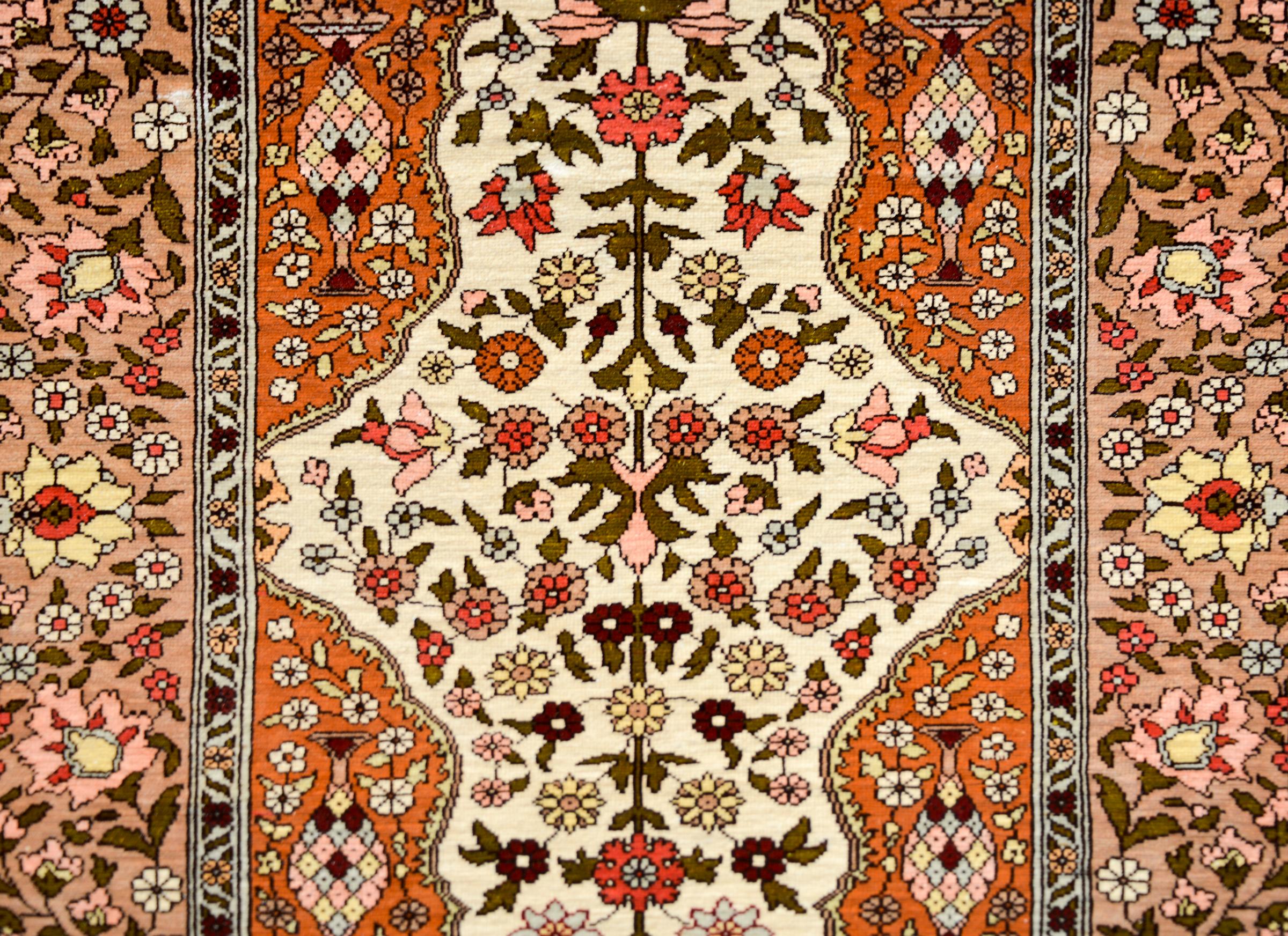 A brilliant vintage silk Turkish Hereke rug woven with a central medallion with a mirrored floral pattern woven in pink, crimson, yellow, and dark green, against a cream colored background, and amidst a field of flowers, and surrounded by a