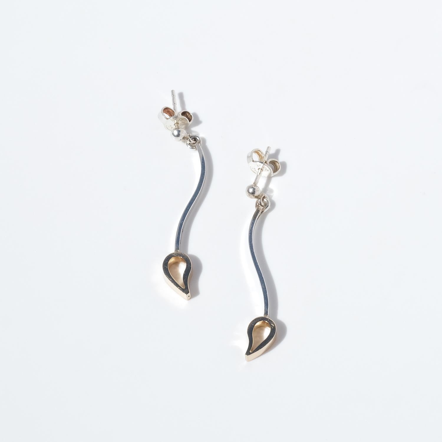 These elegant stud earrings combine sterling silver with 18k gold in a graceful, modern design. Each earring features a slender, wave-shaped silver stem that adds a sense of organic movement. At the end of the stem hangs a three-dimensional gold