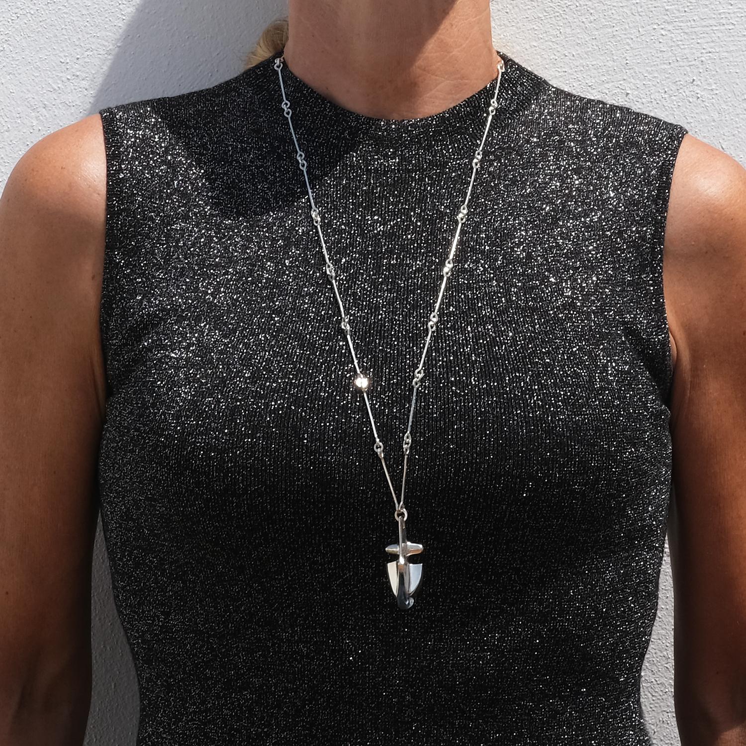 This sculptural necklace in silver and blue acrylic is titled Iguana, a name that brings to mind the graceful curve and crest-like form of the reptile. The pendant features a bold, asymmetrical silver frame that gently arcs around a deep blue,