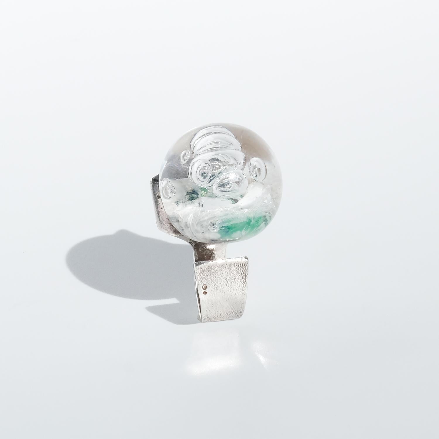 This vintage silver ring, called Frozen Lake, features a striking, wide band with an open design that gives it a sculptural, airy quality. Its round acrylic centerpiece, infused with swirling blue and green pigments and delicate air bubbles,
