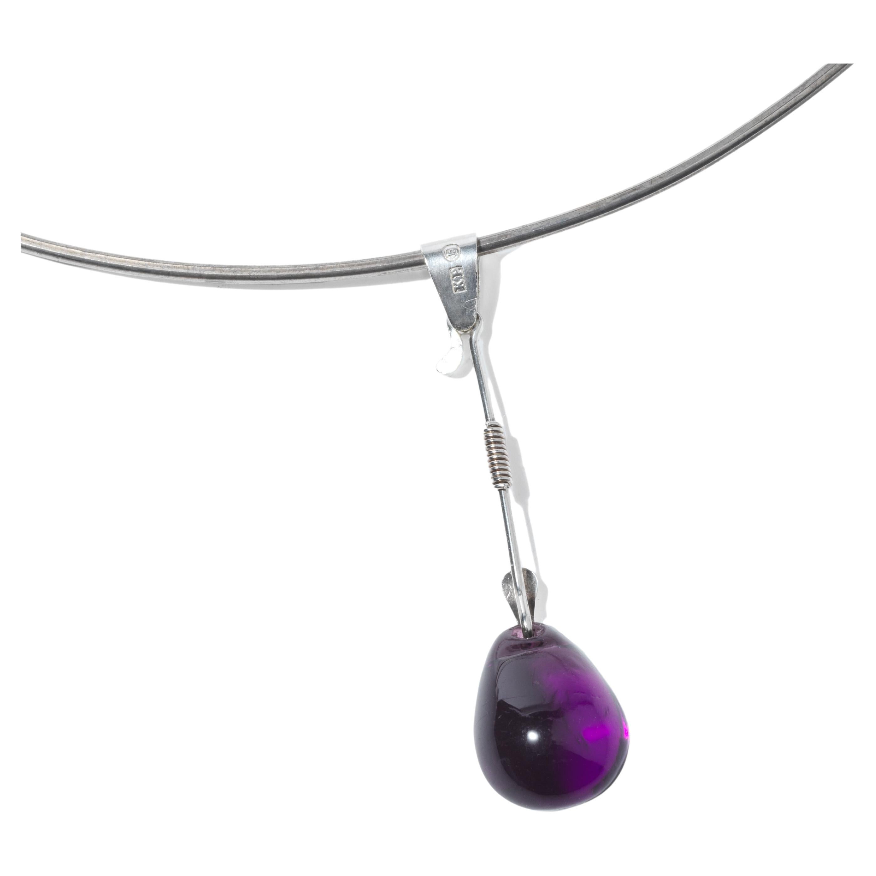 This neck ring is a simple, elegant silver piece with a sleek and modern design. A single, teardrop-shaped amethyst pendant dangles gracefully from the center, its deep purple hue providing a striking contrast against the silver. When worn, the neck