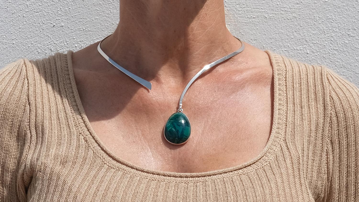 This sculptural necklace features a sleek silver neck ring with a soft, asymmetrical form—gently tapering on one end and widening slightly at the open side. Suspended from it is a teardrop-shaped pendant set with a cabochon-cut Eilat stone, known