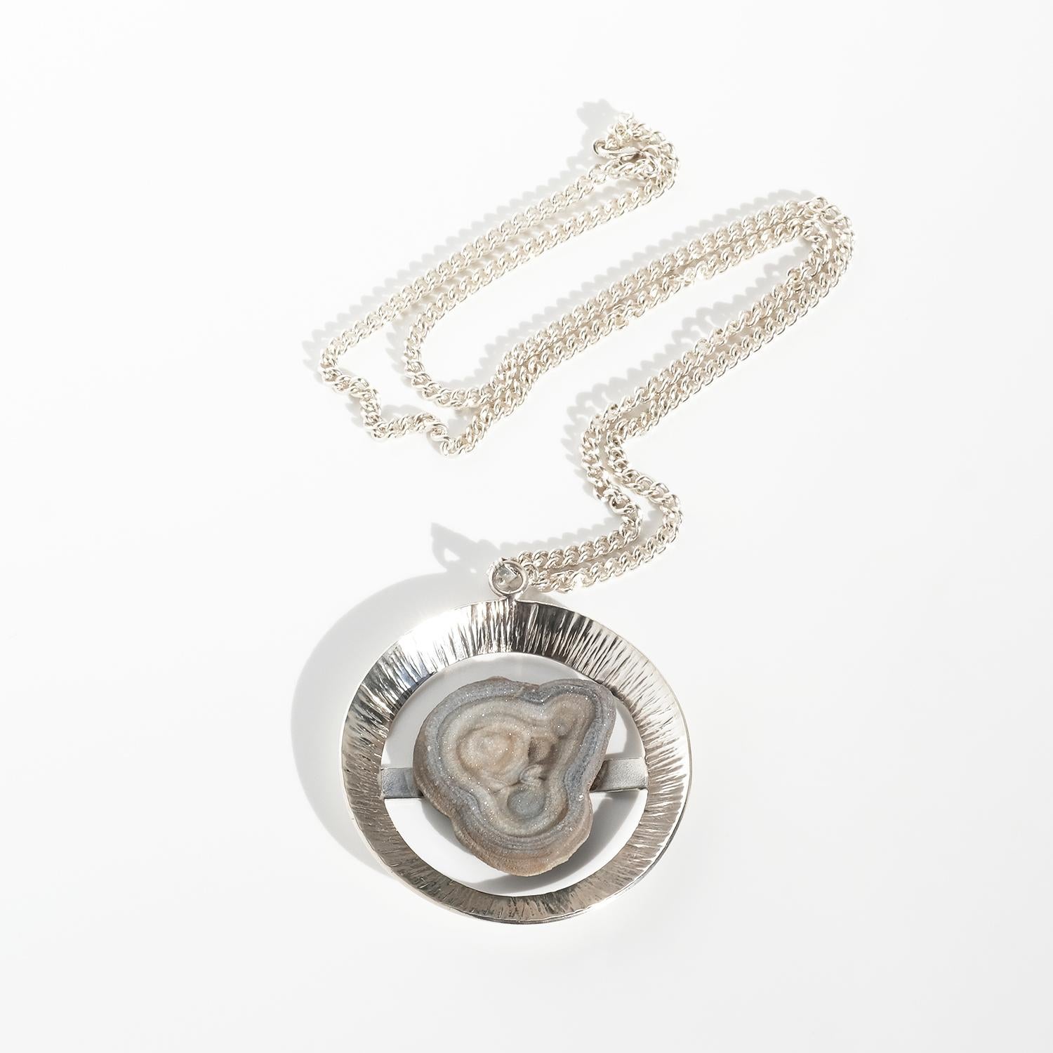 This striking pendant is crafted in silver and set with a natural fossil. It features a broad circular silver frame with a textured, radiating surface that catches the light. At its center, a slice of fossil stone is held by a horizontal silver bar.