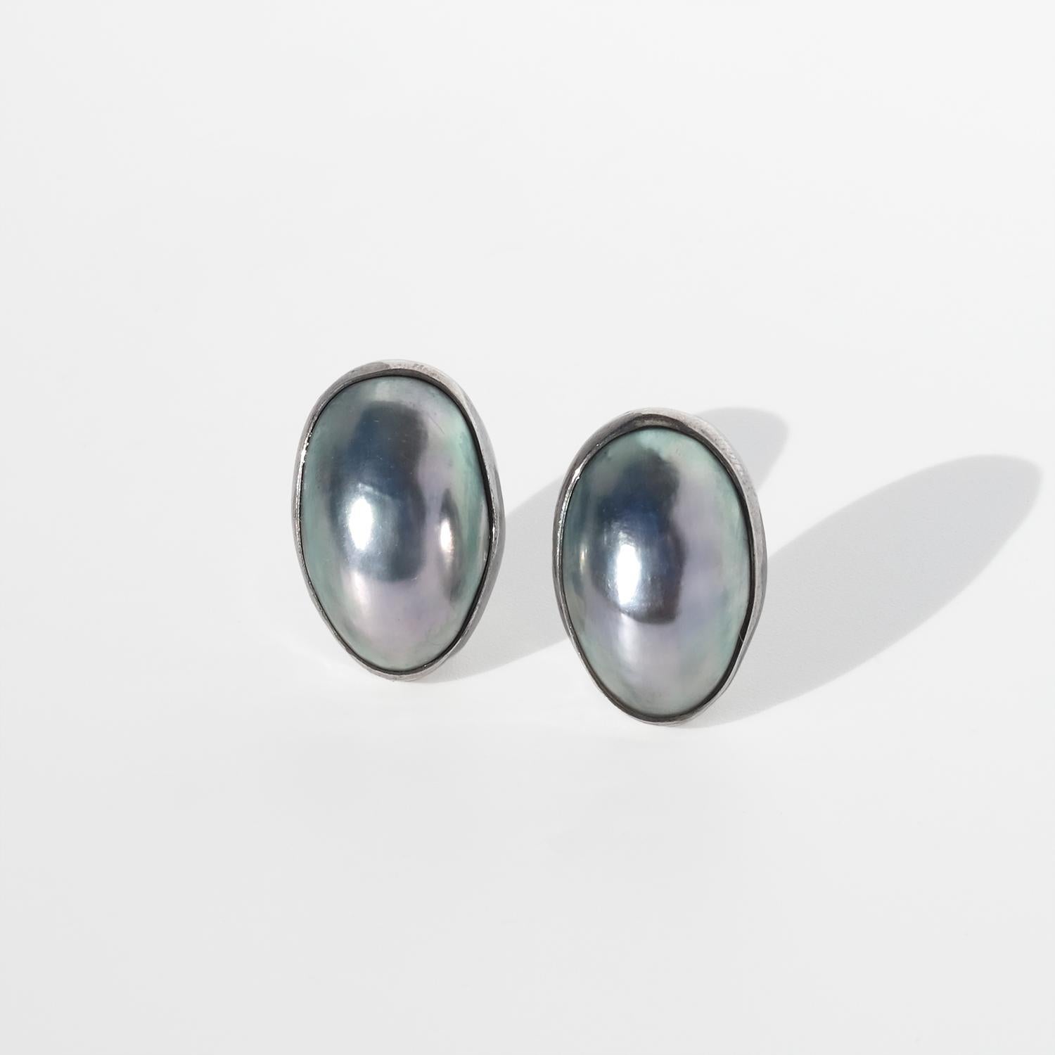 Questi orecchini vintage in argento presentano grandi perle ovali cabochon di madreperla grigia incastonate in castoni lisci e sagomati che seguono il contorno organico di ogni perla. Le superfici mostrano un'iridescenza morbida e fredda con toni