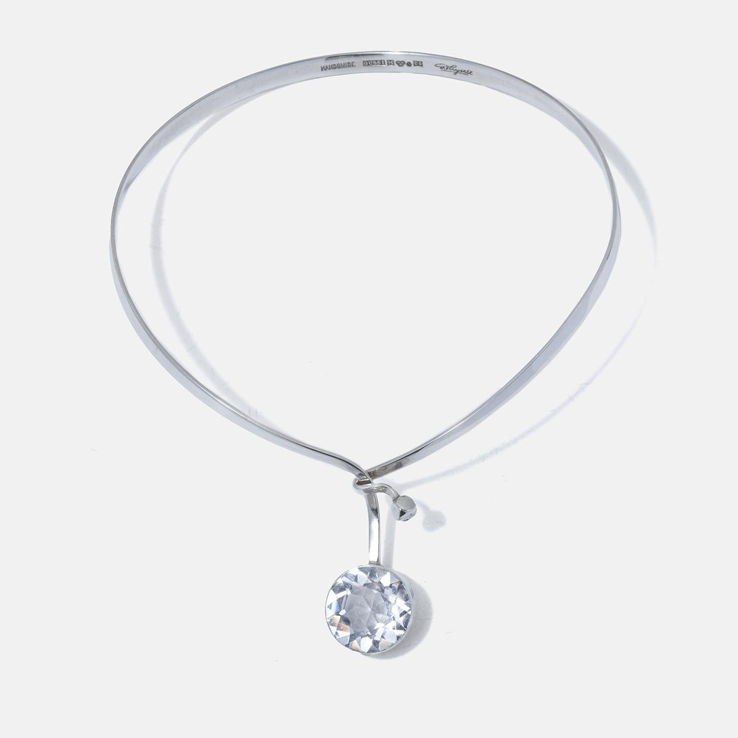 This neck ring is a sleek, silver neck ring that encircles the wearer's neck with a clean and modern design. It has a rigid, collar-like shape that opens at the front, allowing it to wrap comfortably around the neck. A single, large, round rock