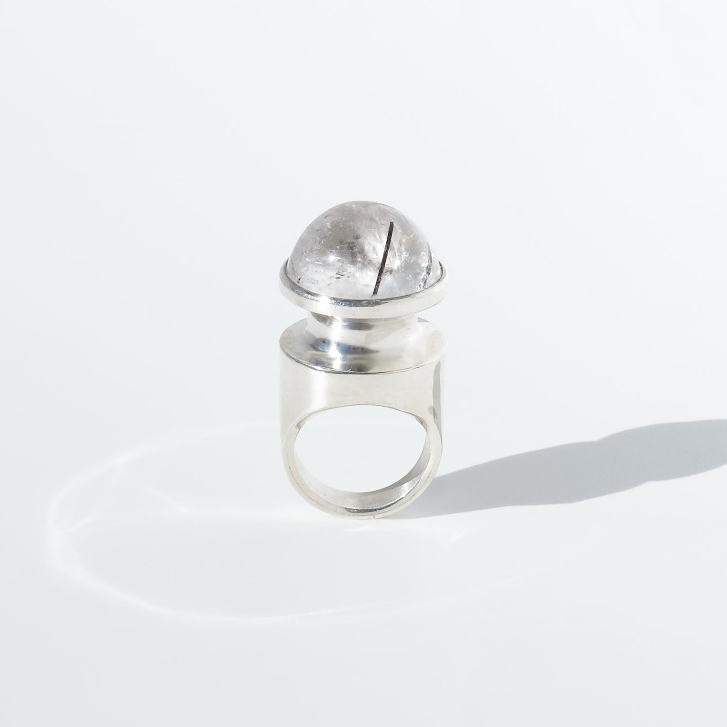 This vintage silver ring features a striking cabochon-cut rock crystal, its smooth, domed surface revealing delicate tourmaline inclusions that create an organic, almost otherworldly effect. The simple bezel setting allows the stone’s natural beauty