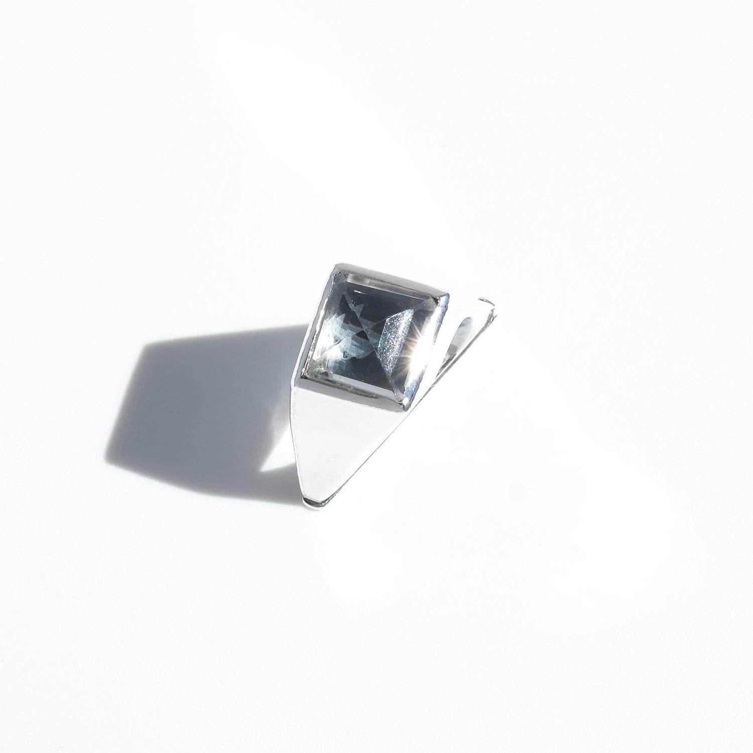 This striking ring is crafted in silver with an asymmetrical design that gives it a sculptural, almost architectural presence. The shank has a distinctive triangular shape, adding a bold geometric element to the piece. At the top, a faceted rock