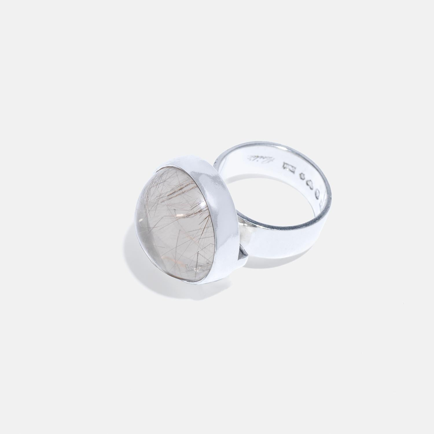 This silver ring features a wide shank and a prominent setting, with a silver bar underneath for added support and decoration. On top, it is adorned with a beautifully encased cabochon-cut rutile quartz stone, showcasing intricate, needle-like