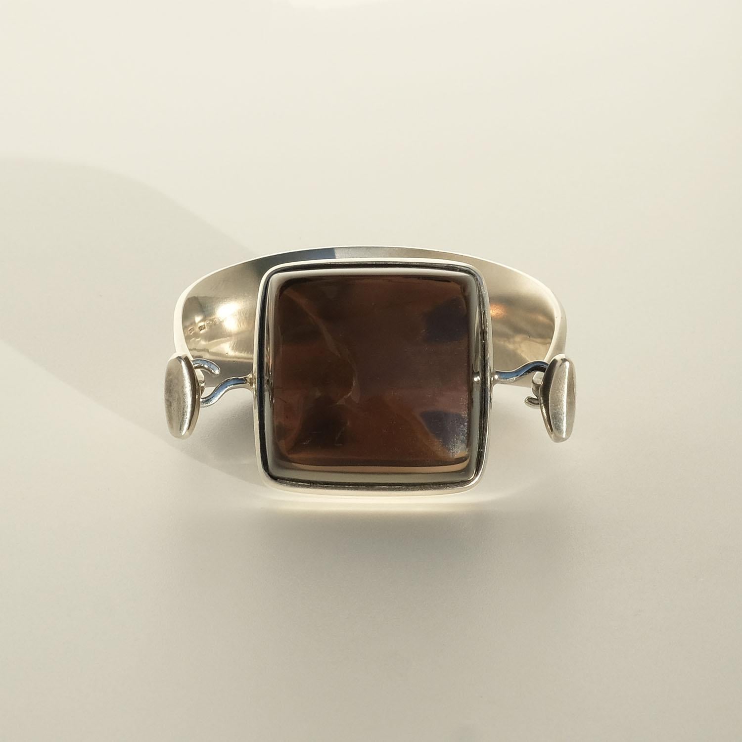 Vivianna Torun Bülow-Hübe (1927-2004) for Georg Jensen (model no 203B). Designed in the 1960s.

This sterling silver fixed bracelet is adorned with a large square smoky quartz stone. The smoky quartz is attached to the bracelet with two hooks of