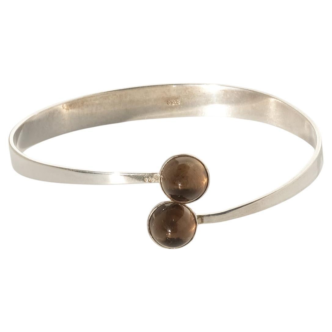 Esta pulsera puño de plata presenta dos gemas de cuarzo ahumado, talladas en estilo cabujón liso, con sus tonos marrones cálidos y translúcidos que añaden una sutil profundidad y elegancia. Las piedras están engastadas en cada extremo del puño