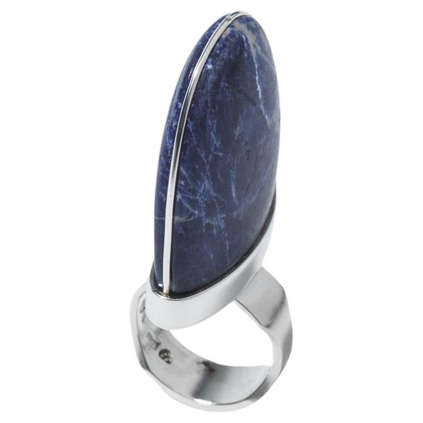Dies ist ein silberner Ring mit einem großen, körnigen blauen Sodalith Stein prominent in der Mitte gesetzt. Der Sodalith-Stein hat einen ausgeprägten Silberstreifen, der sich durch seine Mitte zieht. Die kühne Fassung und der unterschiedlich breite