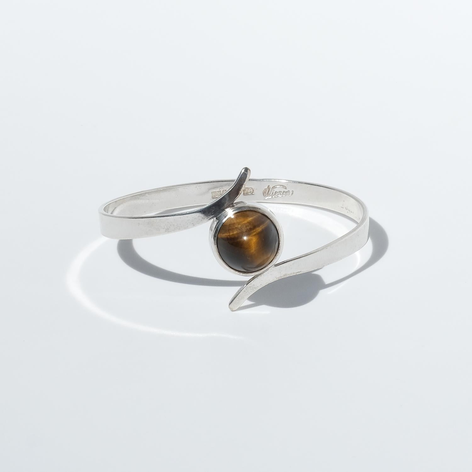 Questo bracciale vintage è realizzato in argento con una struttura solida e fissa. Presenta una gemma occhio di tigre con taglio cabochon incastonata in un castone rotondo e liscio. La pietra ha una superficie a cupola lucidata con le