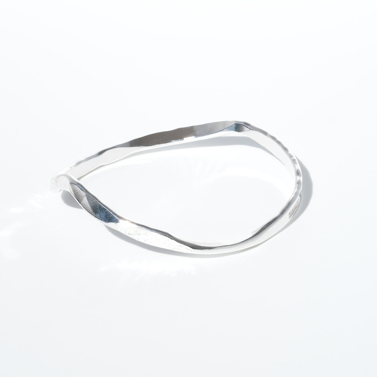Questo braccialetto bangle in argento vintage ha una forma asimmetrica e organica con dolci torsioni e curve irregolari. La sua superficie presenta un mix di texture lisce e sottilmente martellate, che le conferiscono una qualità tattile forgiata a