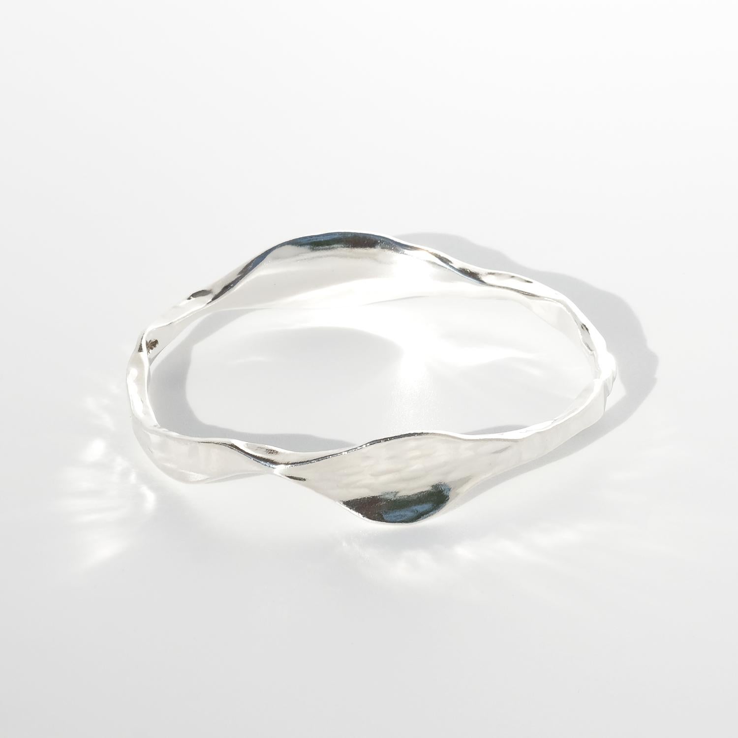 Esta pulsera brazalete de plata presenta una forma elegantemente irregular y orgánica que fluye con naturalidad alrededor de la muñeca. Su superficie está sutilmente texturizada, creando un hermoso juego de luces y sombras sobre la plata pulida y