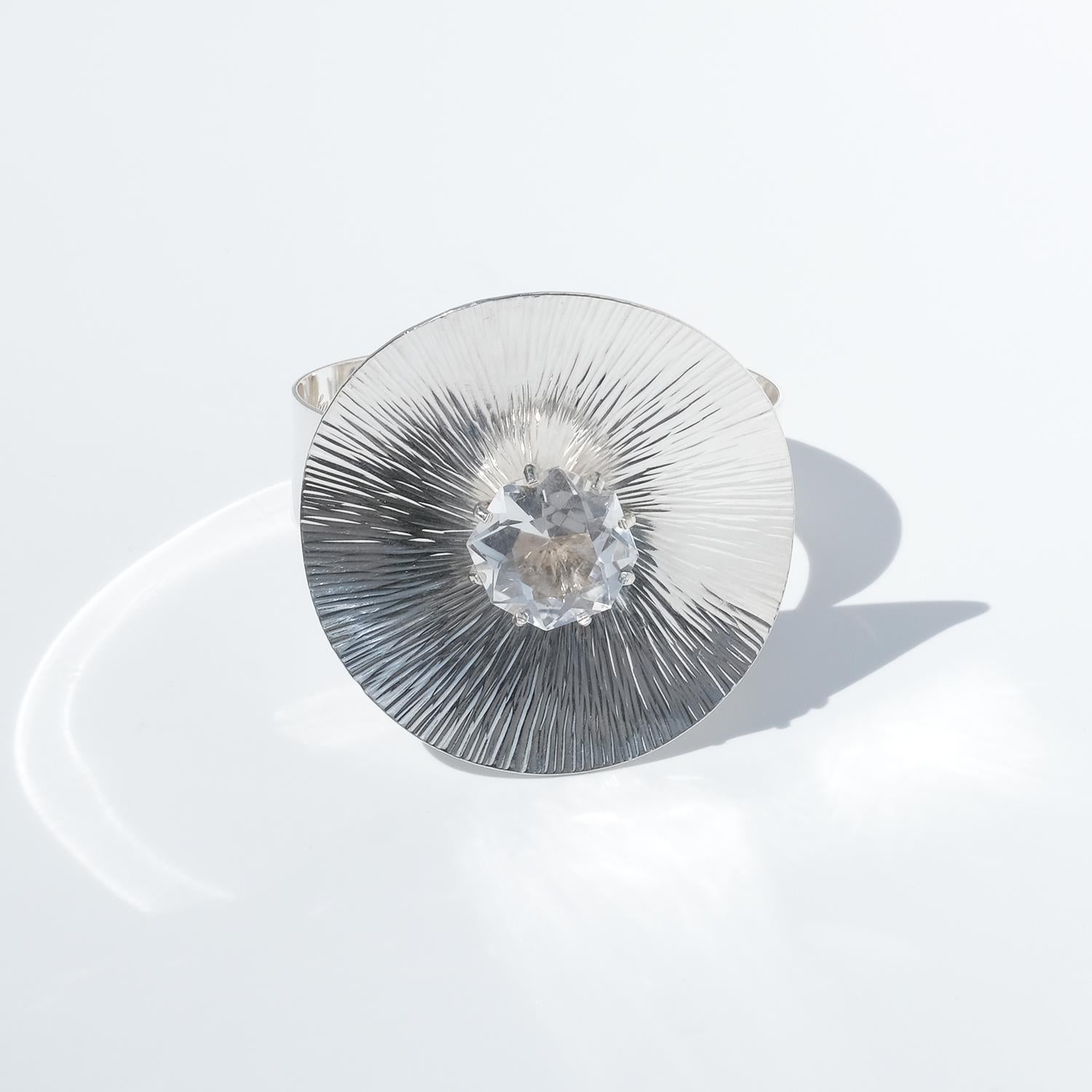 Bracelet vintage en argent orné d'un grand cristal de roche rond maintenu par des griffes en relief. Le cristal est serti au centre d'un large disque concave dont la forme rappelle celle d'un parasol. La surface du disque est finement texturée avec