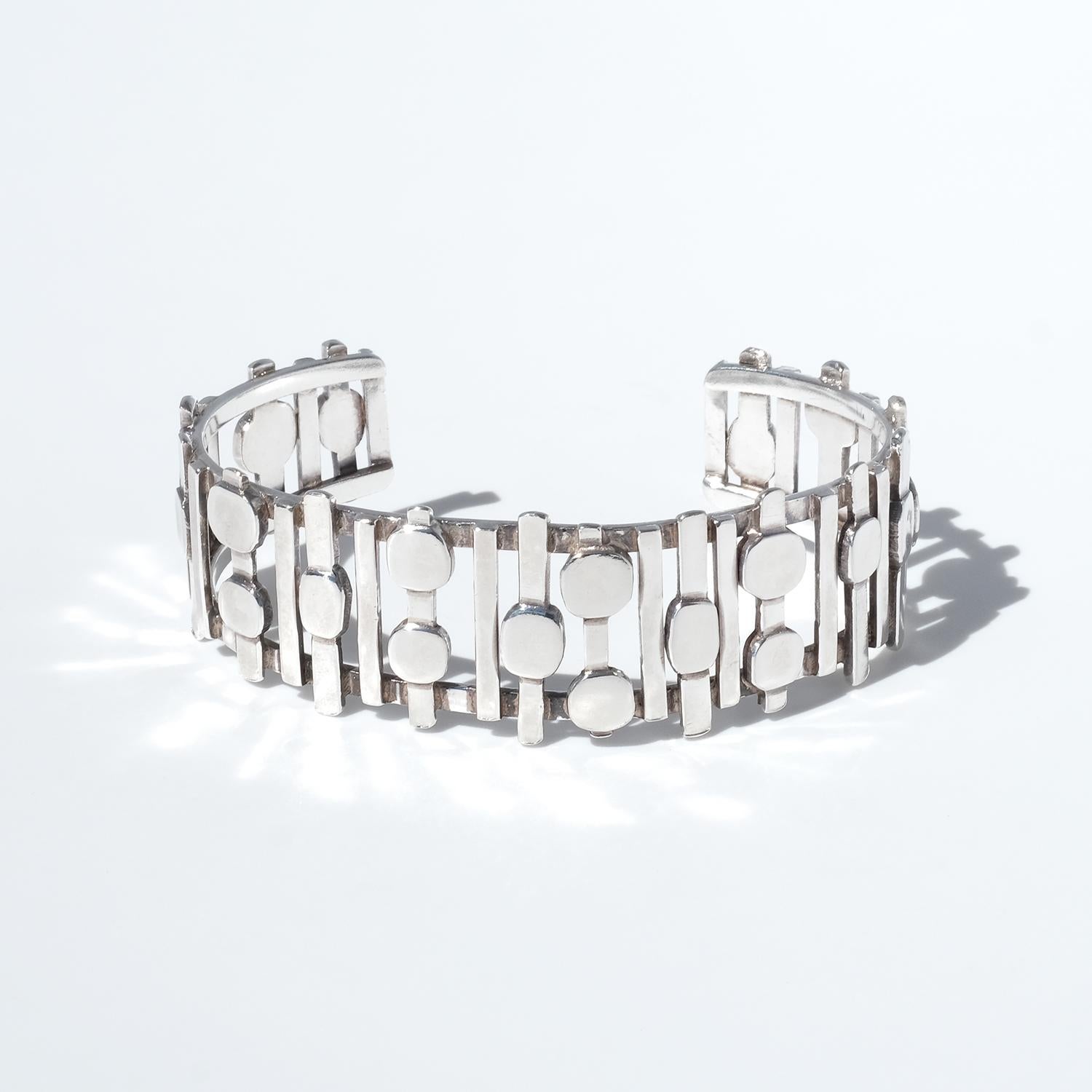 Questo bracciale vintage in argento ha un design strutturato con un motivo traforato composto da barre verticali intersecate da forme rotonde e ovali. La disposizione geometrica crea profondità e senso di movimento, conferendo al pezzo un'estetica