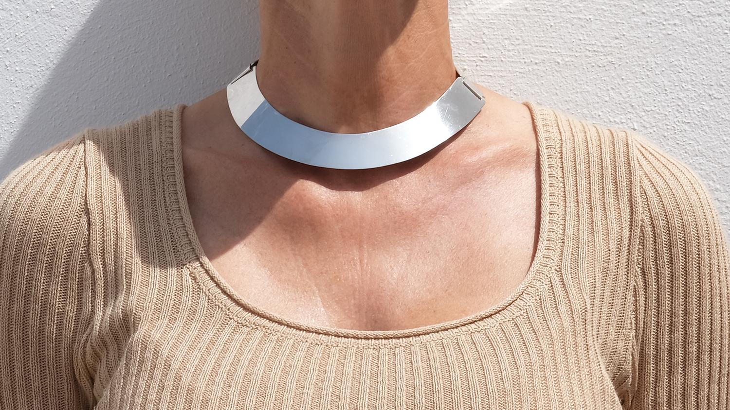 This sculptural silver neck ring features a bold, modernist form that widens gradually toward the front and narrows at the back. Crafted from smooth, mirror-polished silver, the ring is composed of three broad, slightly curved segments joined by