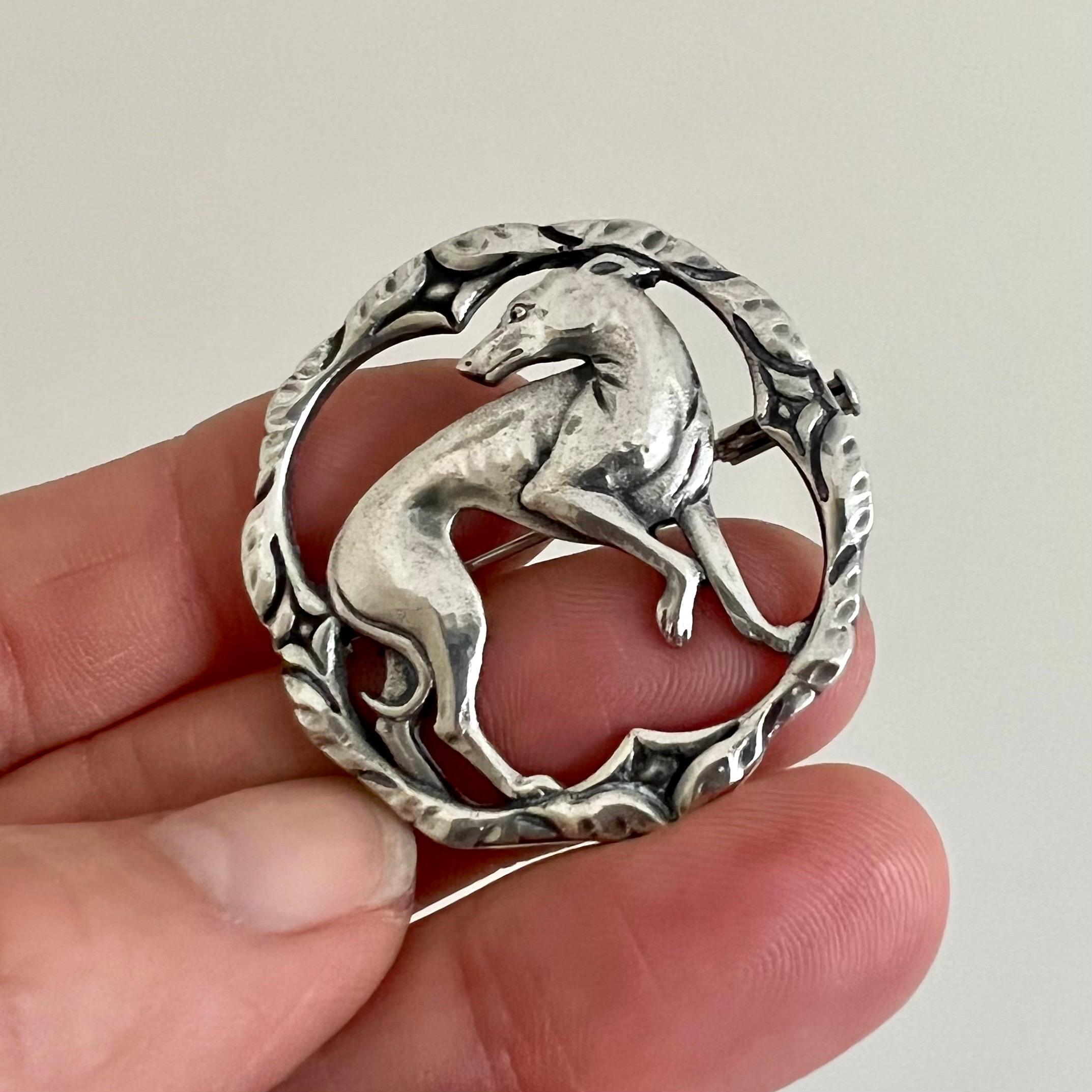 A lovely vintage silver round brooch depicting a greyhound whippet in the middle. The brooch is made of polished silver and beautifully stylized, while the line engravings of the border and the gorgeous dog have a natural patina. At the back of the