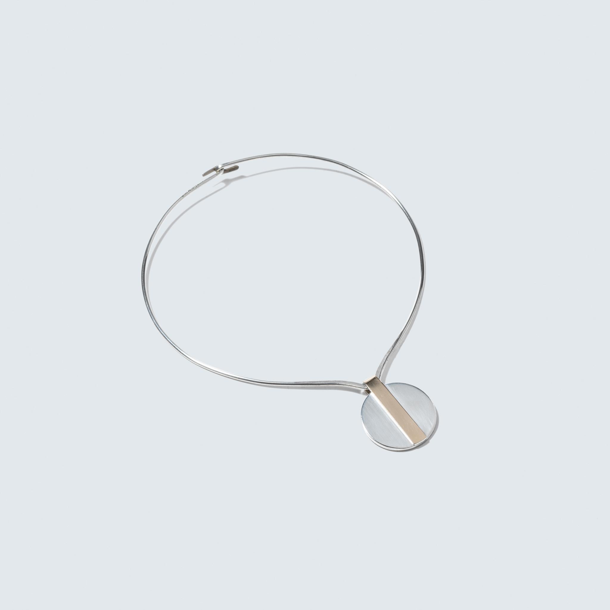 This is a sleek and modern sterling silver neck ring that has a minimalist design. It features a circular silver pendant, which is elegantly simple, with a striking gilded silver strip crossing through its center, adding an authentic touch of
